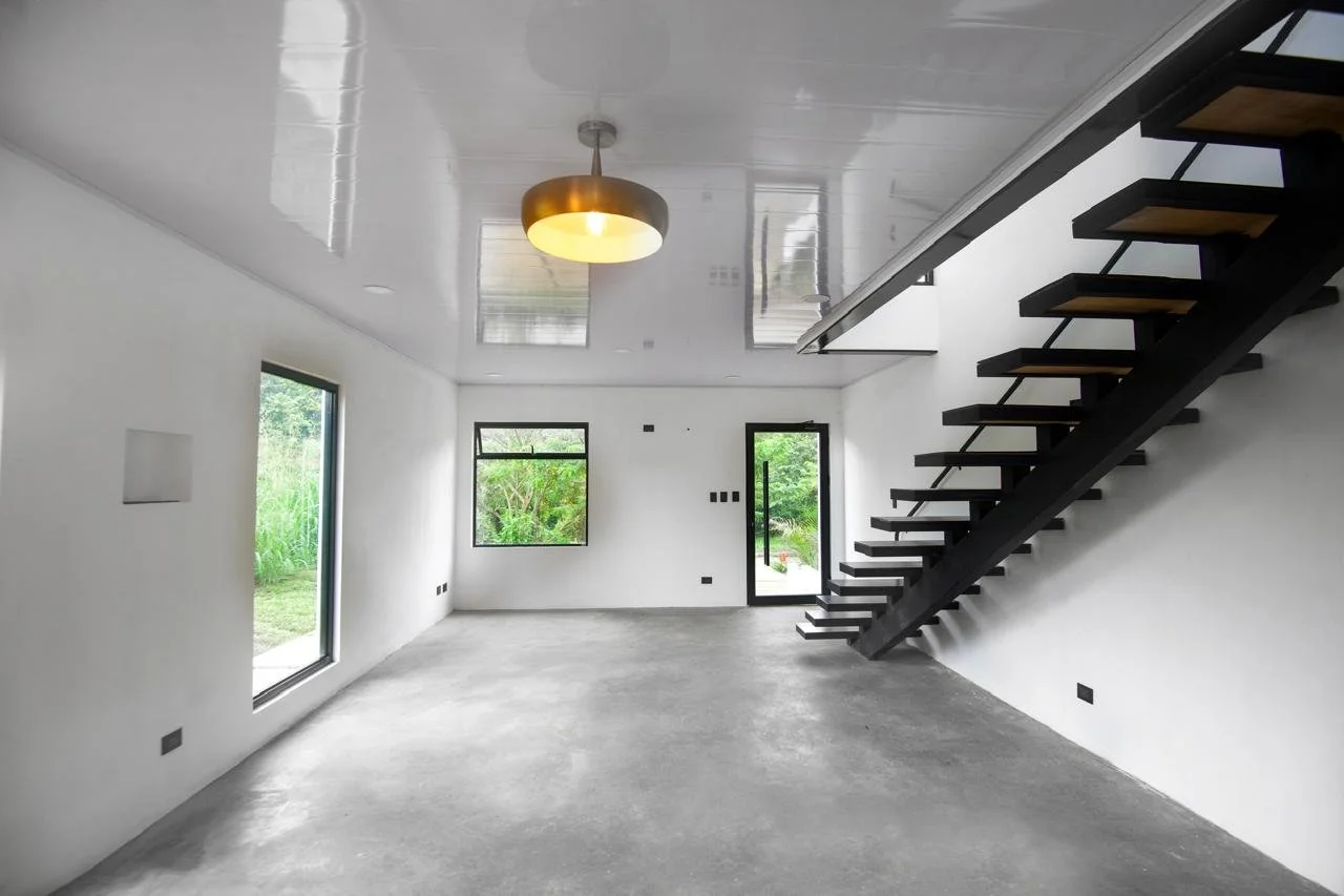Empty living room with large windows, a modern staircase with black steps and wooden treads, white walls, a white ceiling with a gold pendant light, and a concrete floor.