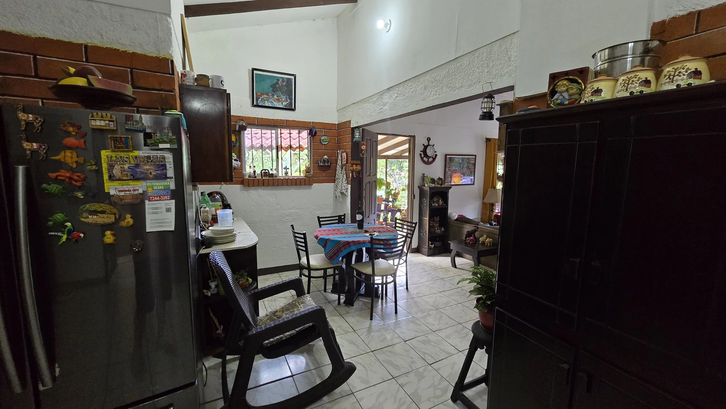 The image shows a cozy kitchen and dining area with a round table covered with a colorful cloth, surrounded by four chairs and a rocking chair nearby. There is a black refrigerator decorated with magnets, a small counter with dishes, and a window wit
