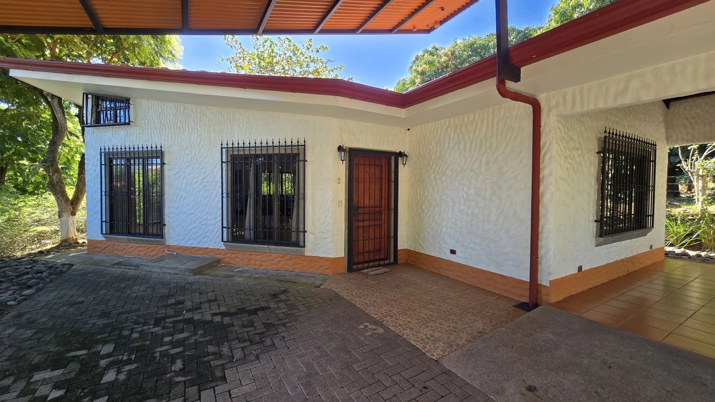 A white house with black window bars, a wooden front door, and a red gutter, surrounded by trees and greenery, with a paved and tiled walkway in front.