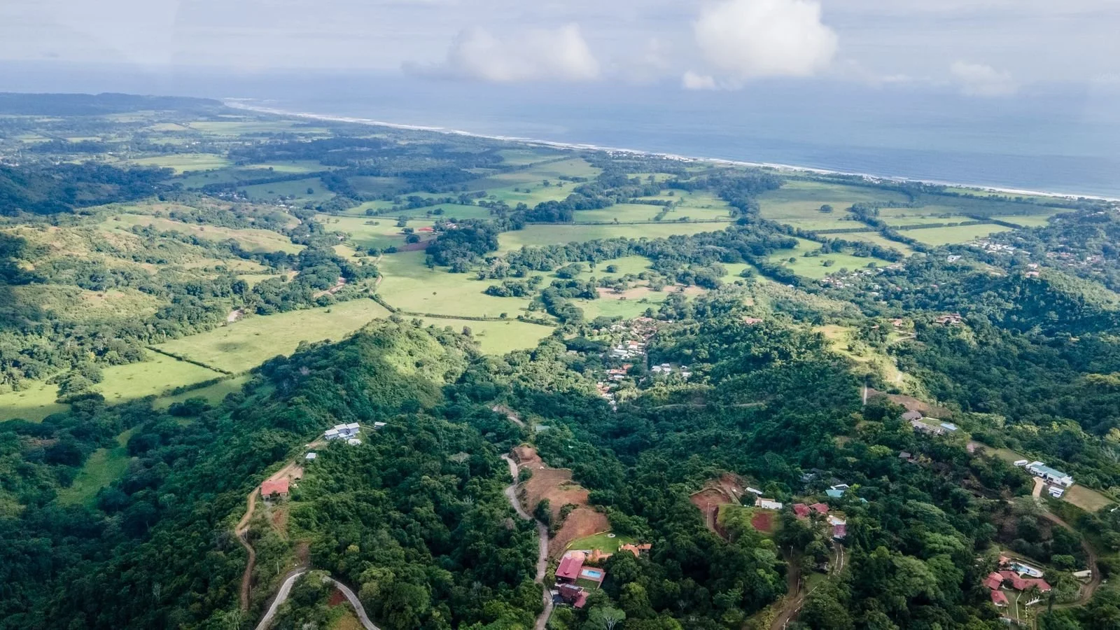 Aerial view of lush green hilly landscape with scattered houses, fields, and trees, extending to a coastline with ocean waves and beaches.