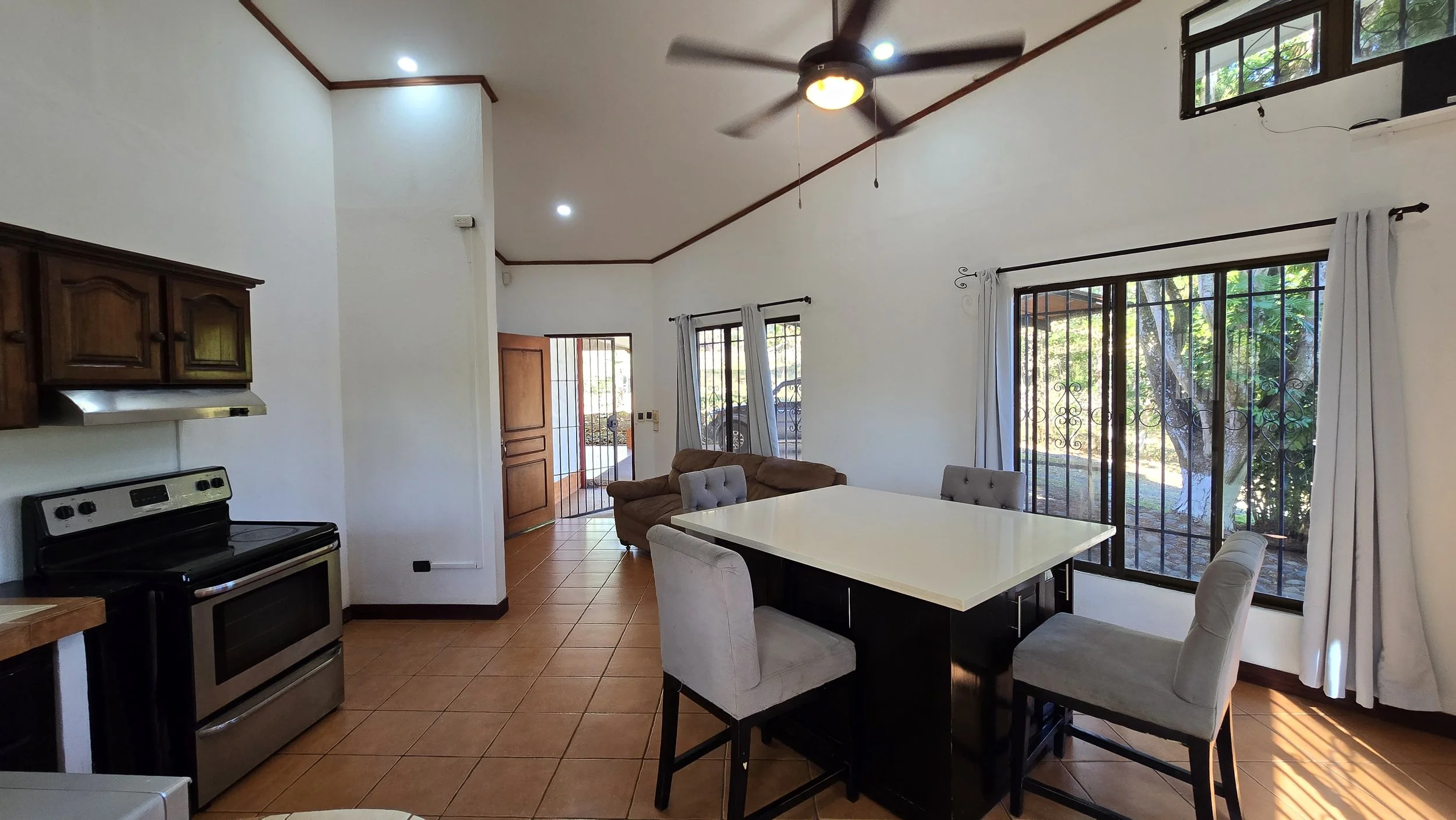 Interior view of a living and dining area with tiled floor, white walls, large windows with bars and curtains, a ceiling fan, a brown sofa, a white dining table with four chairs, and a doorway leading outside.