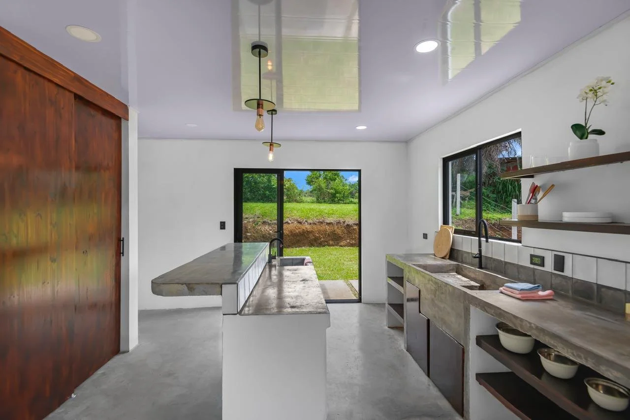 Modern kitchen with concrete countertops, open shelves with bowls and plants, black sink, sliding glass door leading to a grassy backyard, minimalist lighting, and a large wooden sliding door.