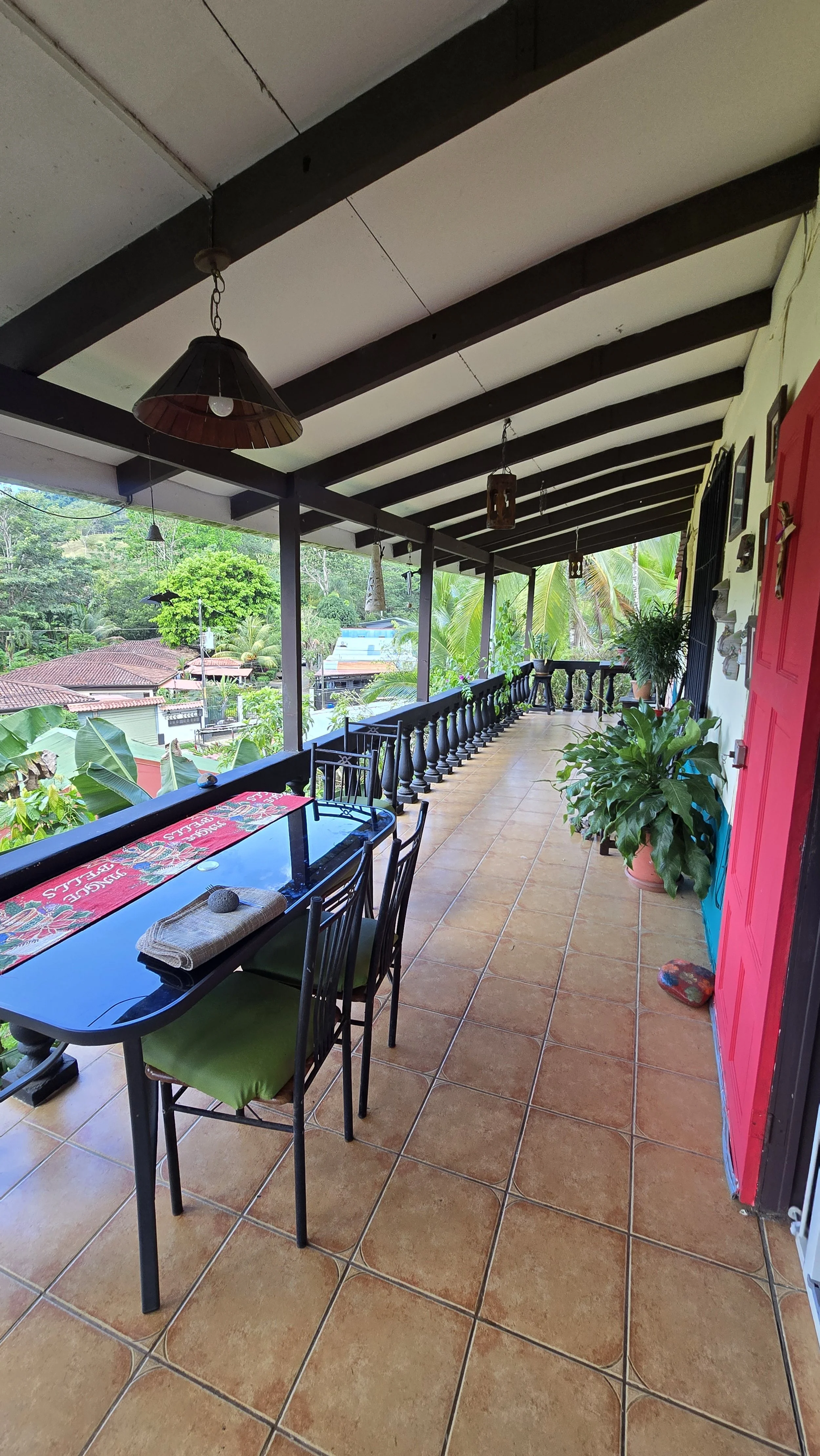 A covered outdoor balcony with tiled floor, black wooden beams on the ceiling, potted plants, a black table with chairs, and a colorful decoration on the wall, overlooking a lush green neighborhood with trees and rooftops.