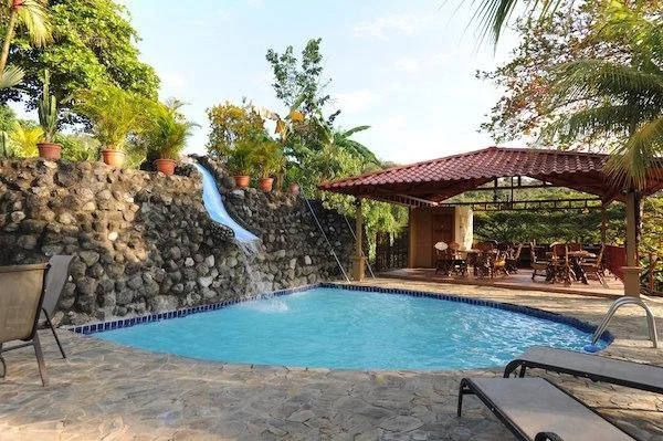 Swim pool with rock waterfall feature and water slide, surrounded by sandy area, outdoor seating area with tables and chairs, and lush greenery including palm trees.