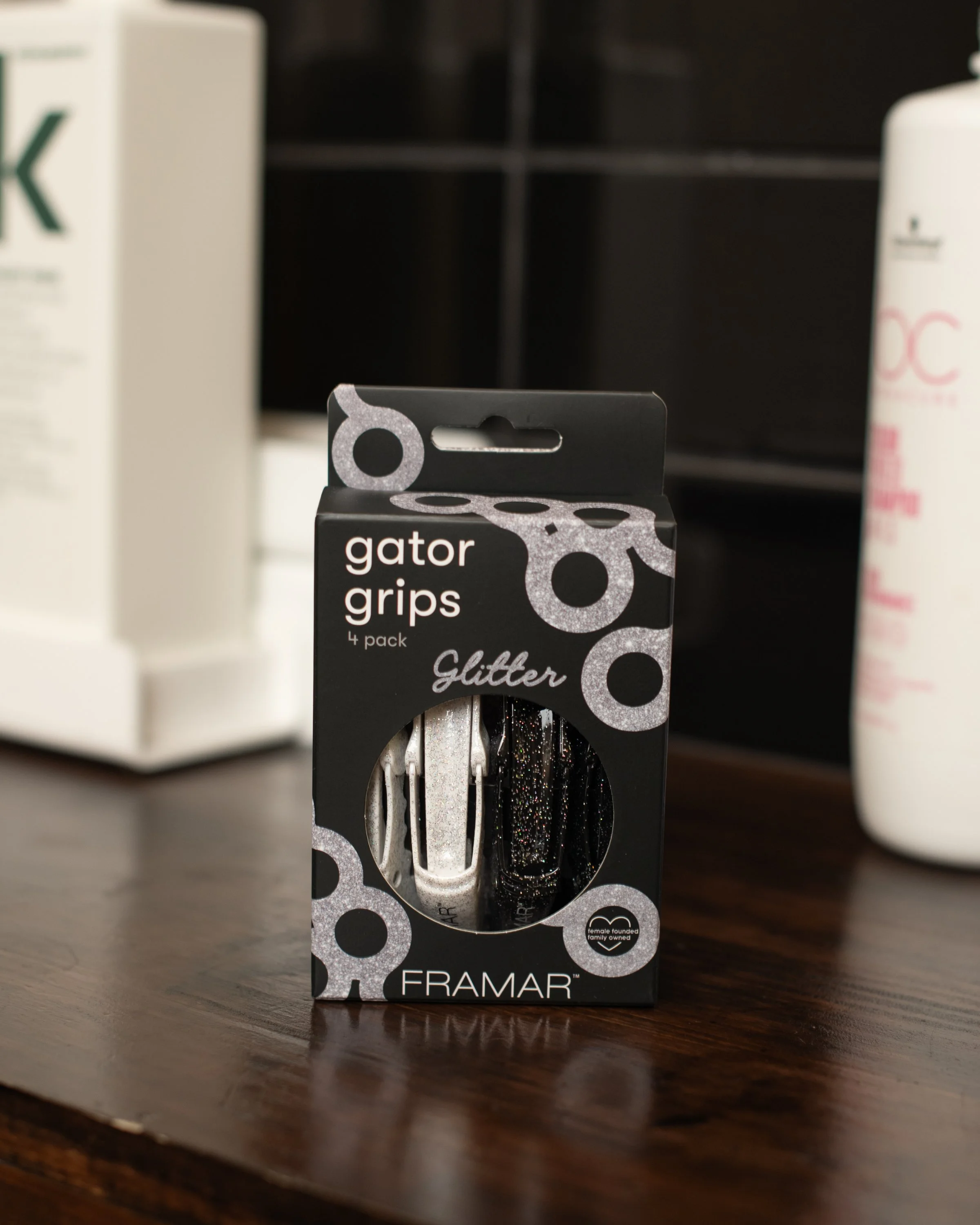 A box of four Gator grips with glitter design, including one in silver and one in black with glitter, placed on a wooden surface with other bottles and packaging in the background.