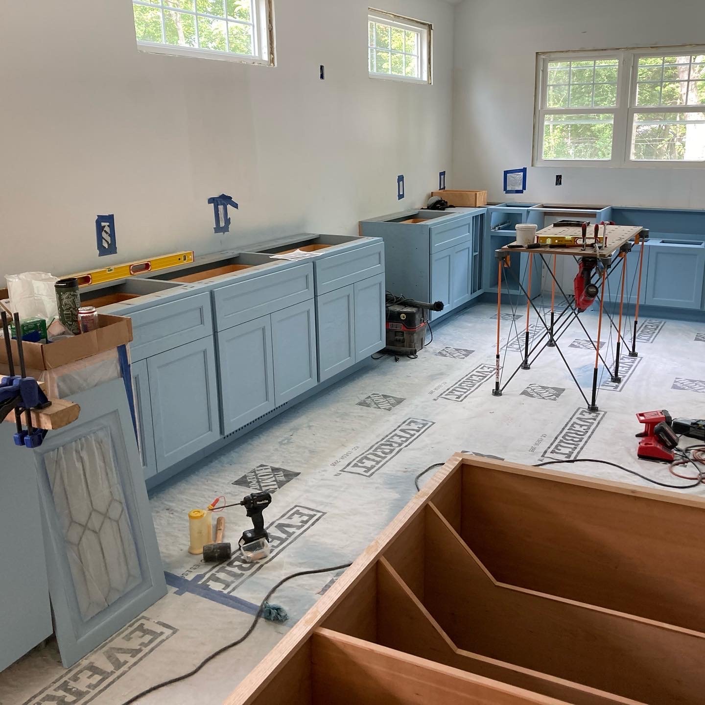Room under construction with light blue cabinets, tools, and equipment. Windows provide natural light. Flooring is protected with material labeled "Everbilt." Some cabinet doors are leaning against the wall.
