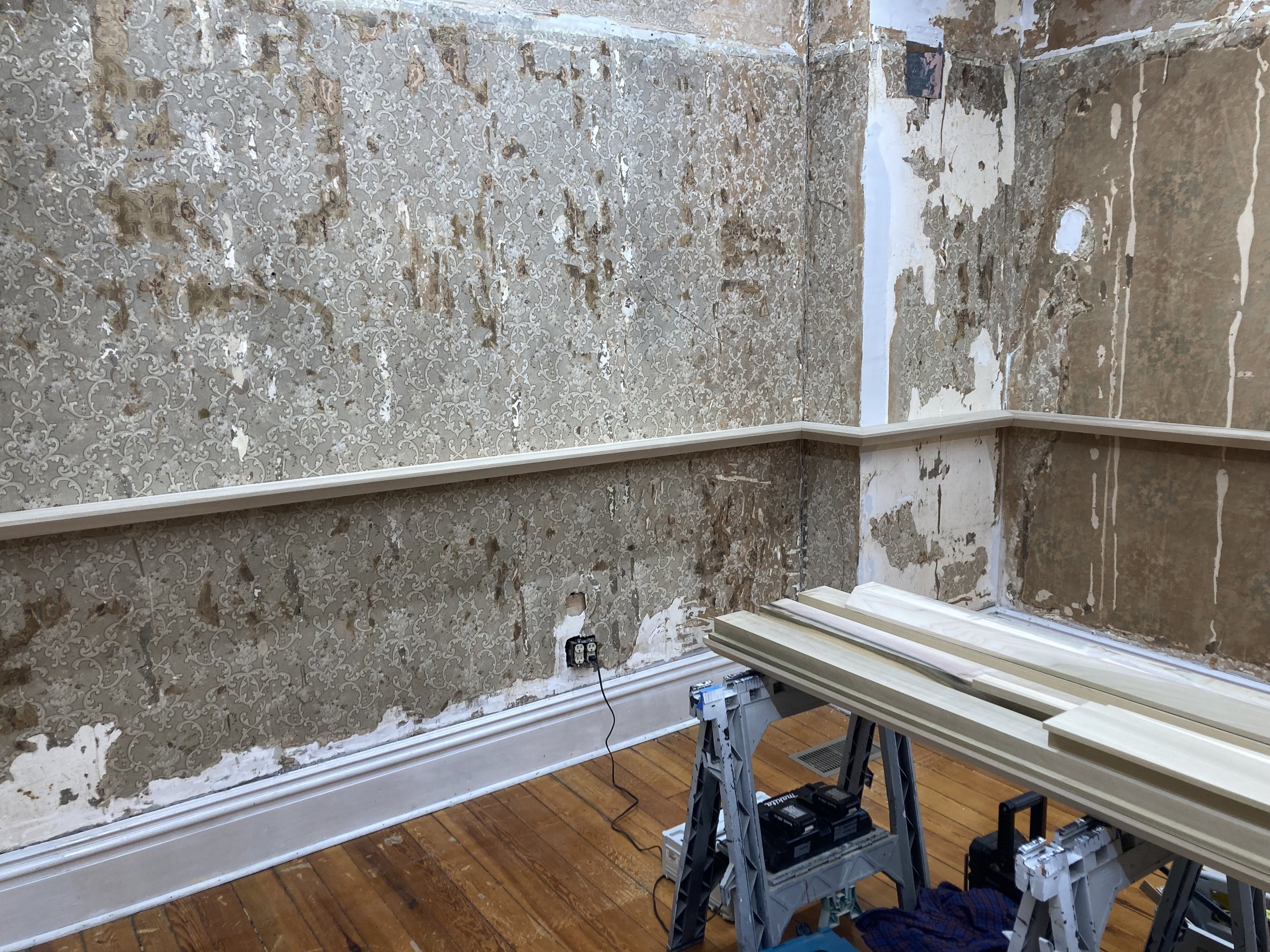 Room under renovation with vintage wallpaper partially removed, exposing plaster walls. Wooden molding and tools are placed on sawhorses in the foreground.