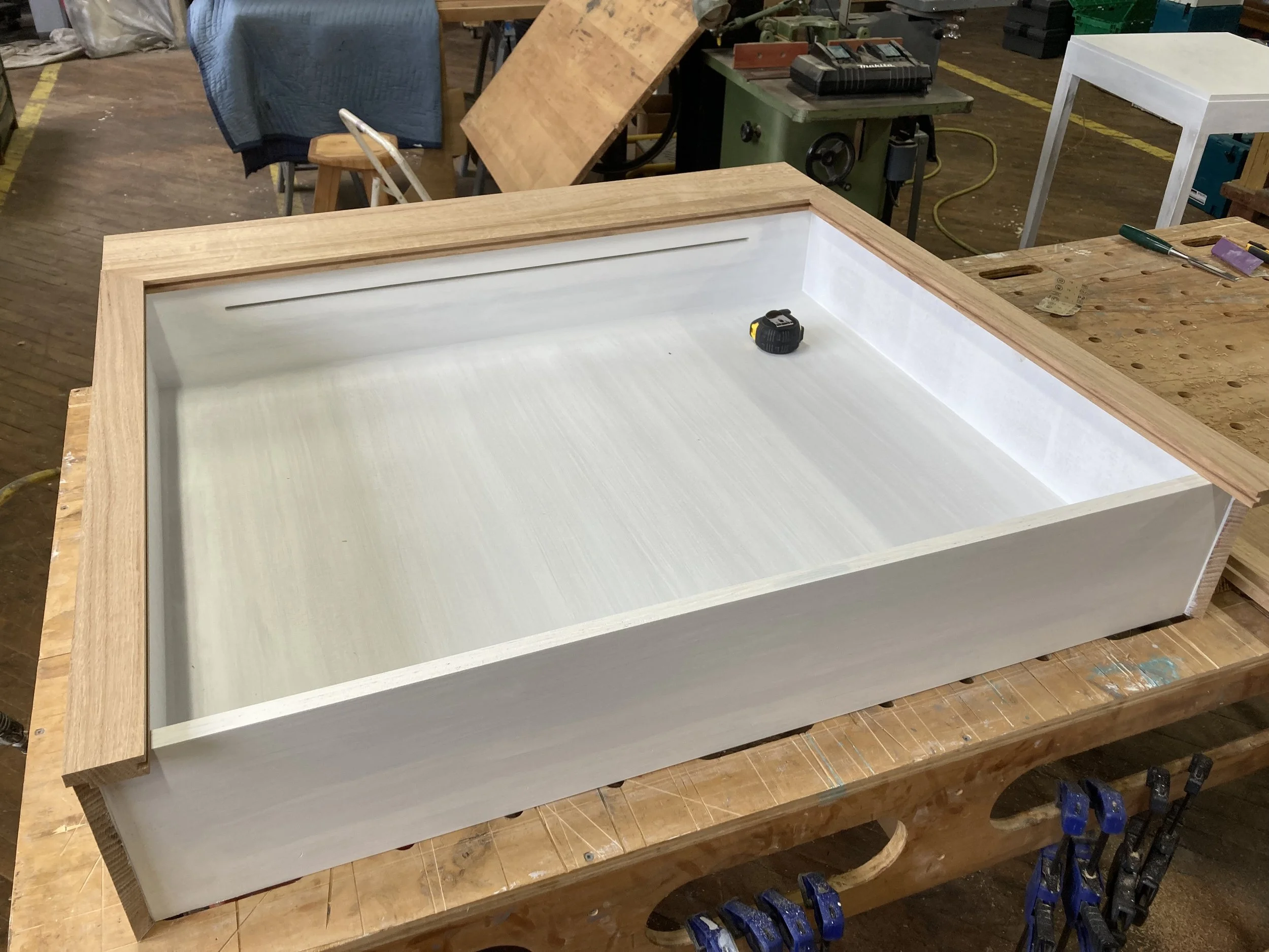 A wooden tray or shallow box on a workbench with a measuring tape inside, surrounded by clamps in a workshop setting.