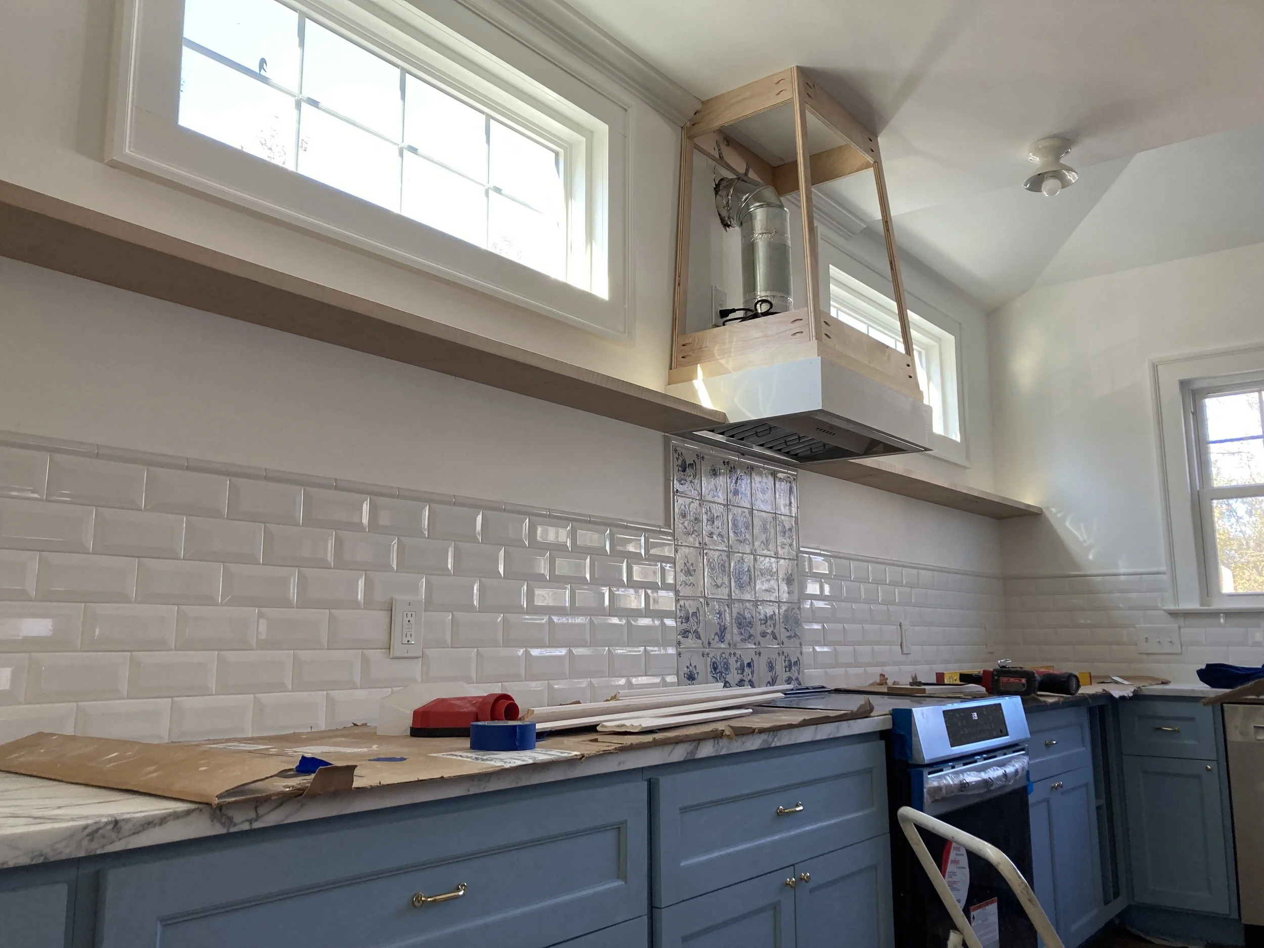 Modern kitchen under renovation with blue cabinets, marble countertops, white subway tile backsplash, and an unfinished range hood. New stovetop and tools visible on countertop.