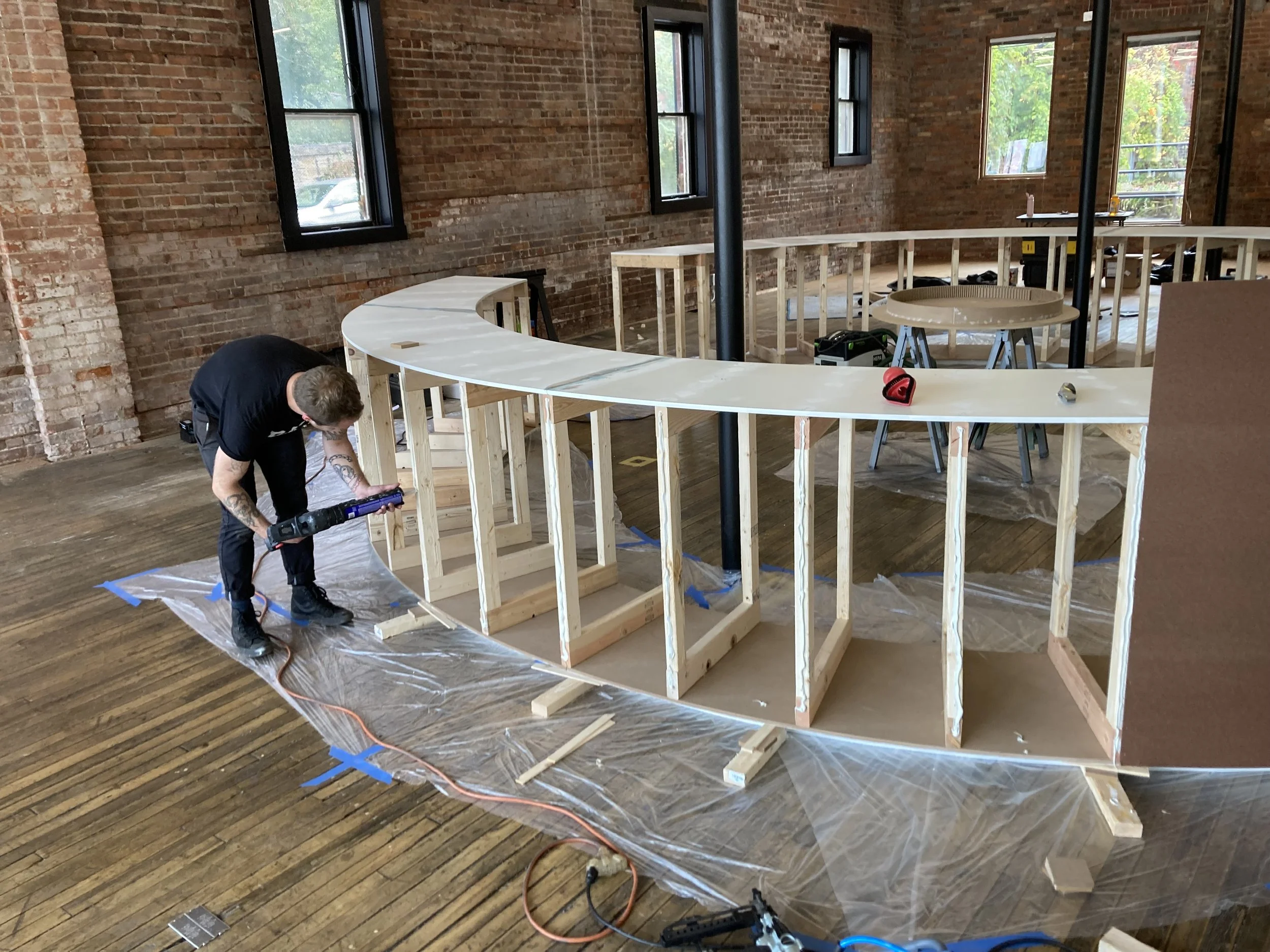 A person working on constructing a curved wooden counter inside a building with exposed brick walls. The person is using a tool, possibly a nail gun or adhesive applicator, on the wooden frame. Plastic sheets and construction materials are visible on