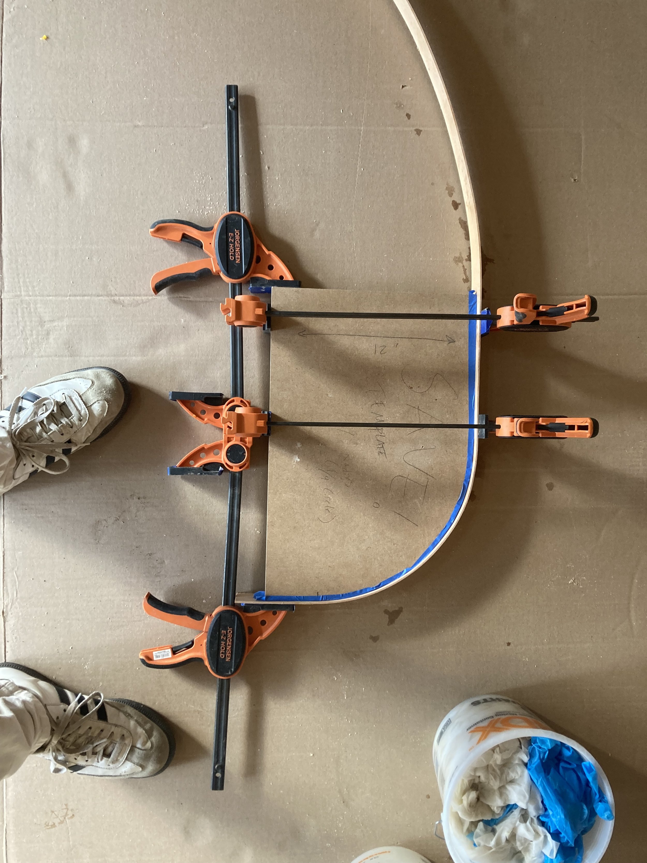 Wood being shaped with clamps, showing an arch in progress, on a cardboard surface with tools and foot in view.