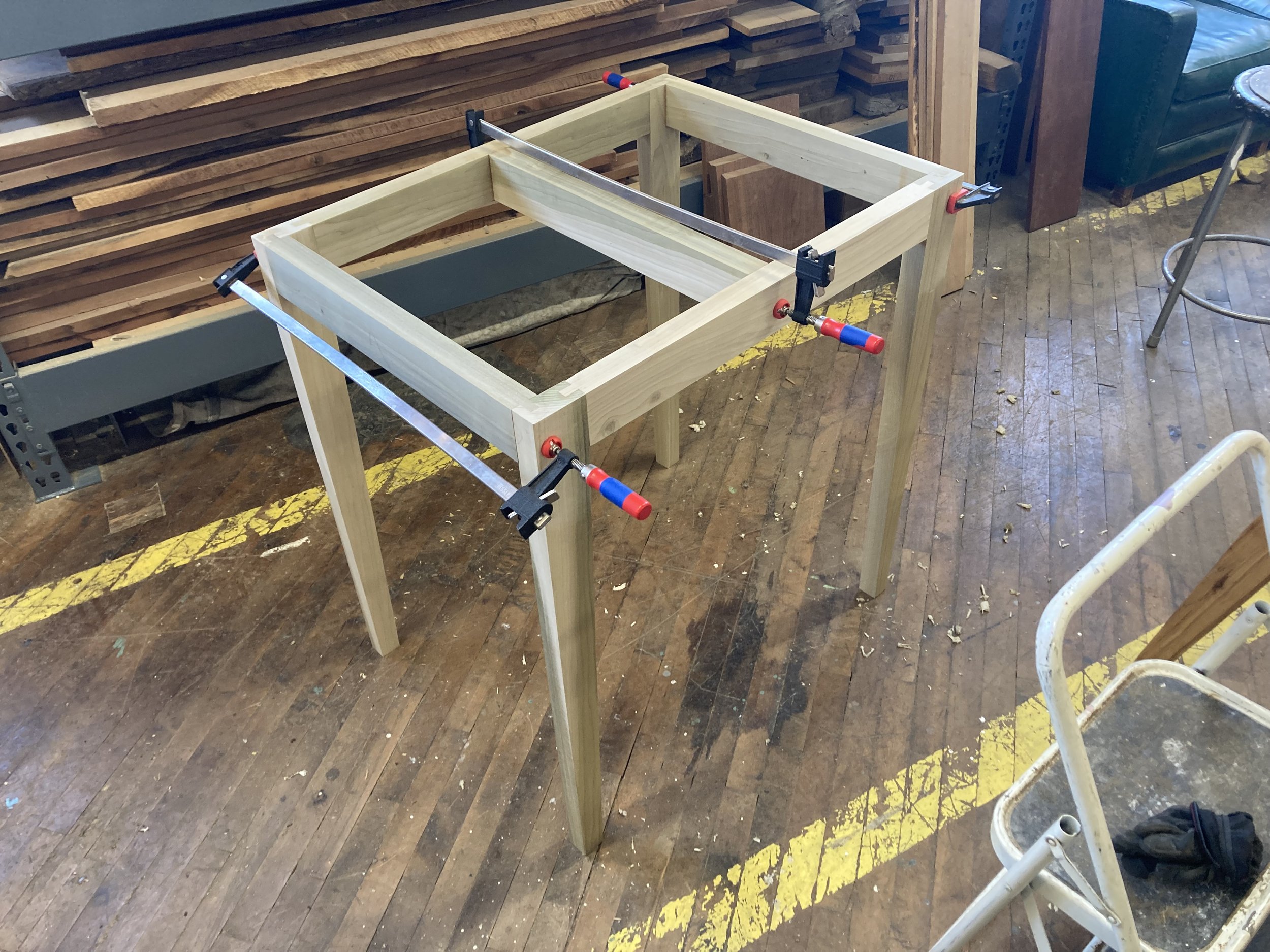 Wooden table frame in a workshop secured with clamps, surrounded by wood planks and tools on a wooden floor.