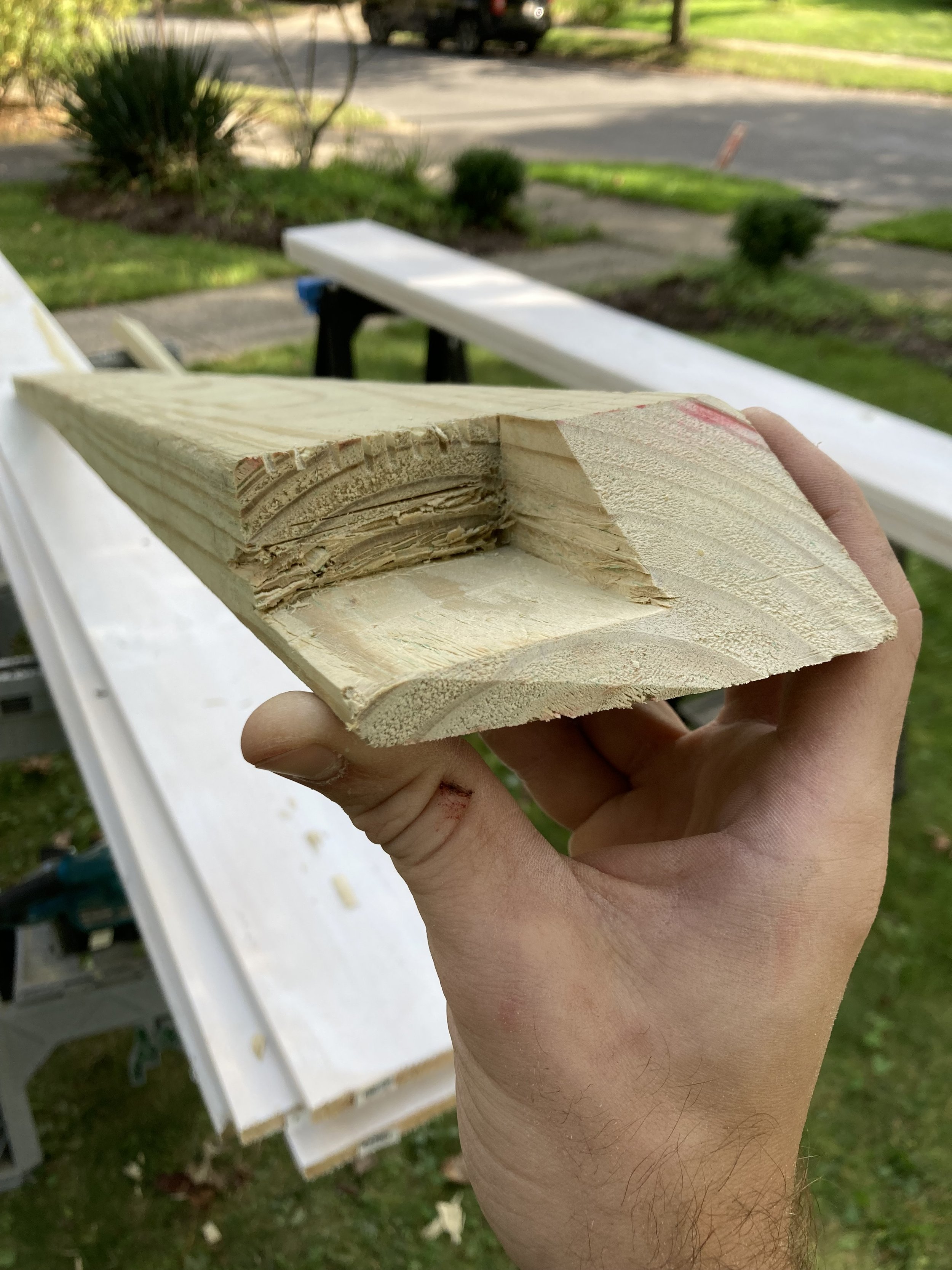 Person holding a piece of wood with a freshly cut groove, outdoors with greenery and woodworking tools in the background.