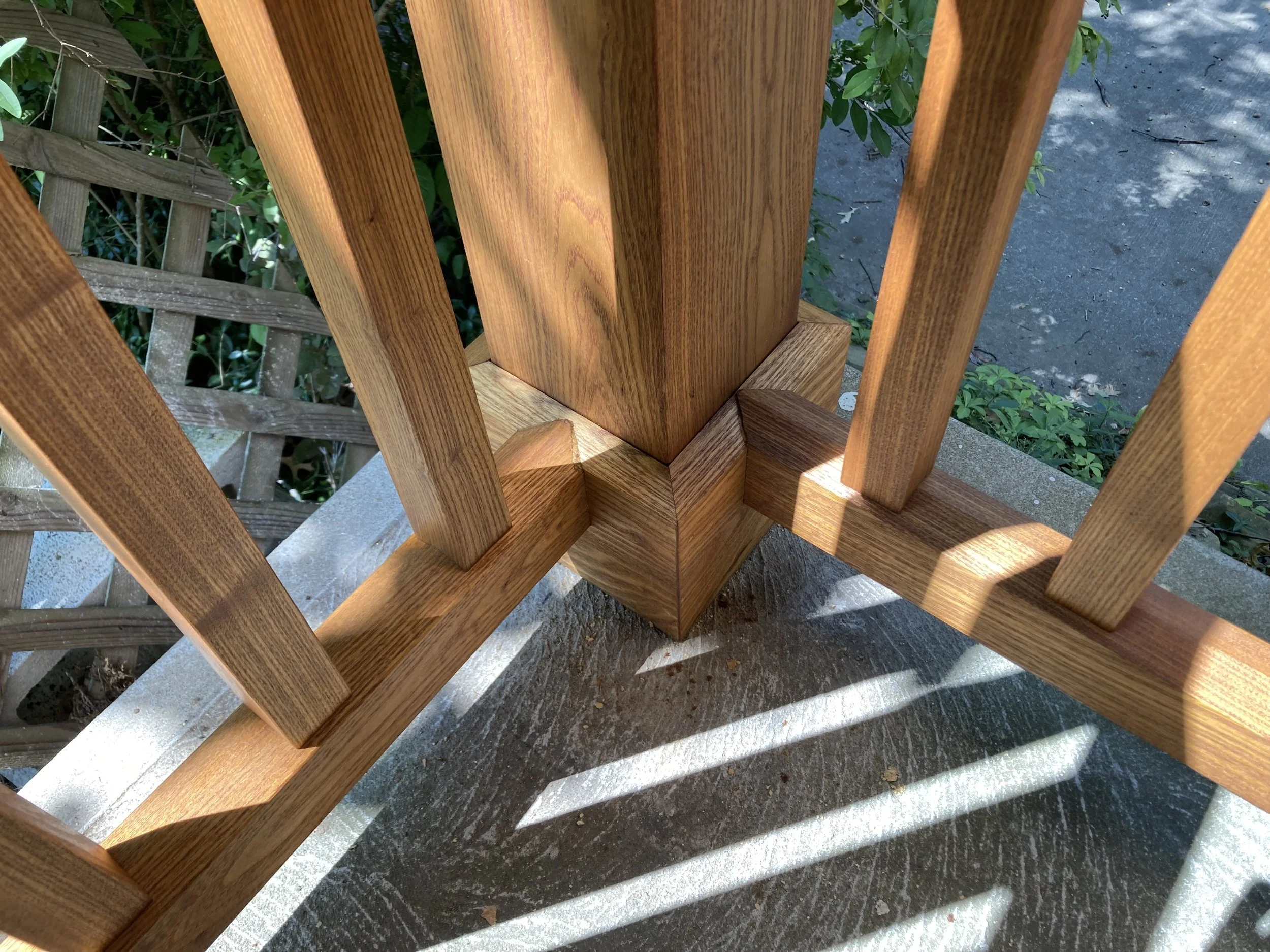 Close-up of a wooden railing corner with vertical and diagonal slats, sunlight casting shadows, and greenery in the background.