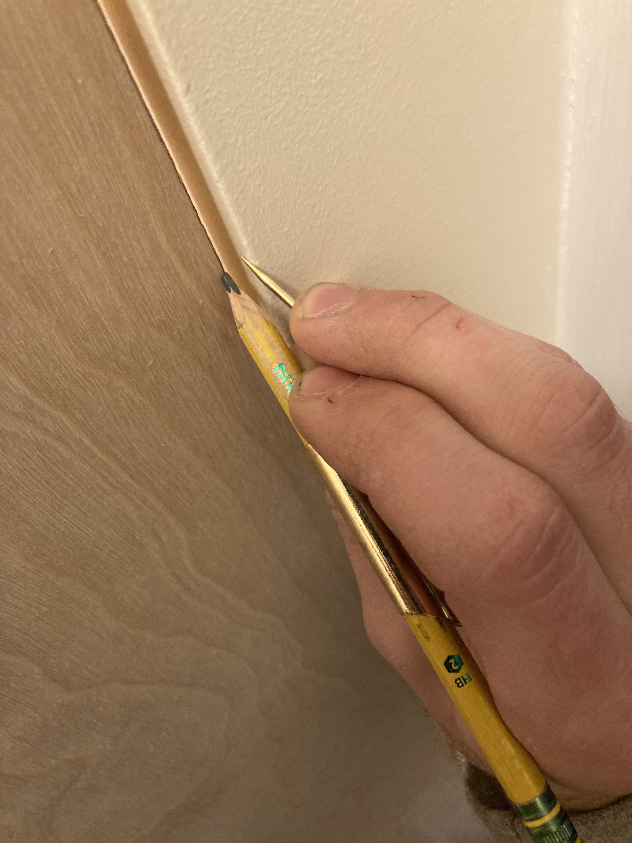Person using a yellow wooden pencil to mark a line along the edge of a wooden surface against a white wall.