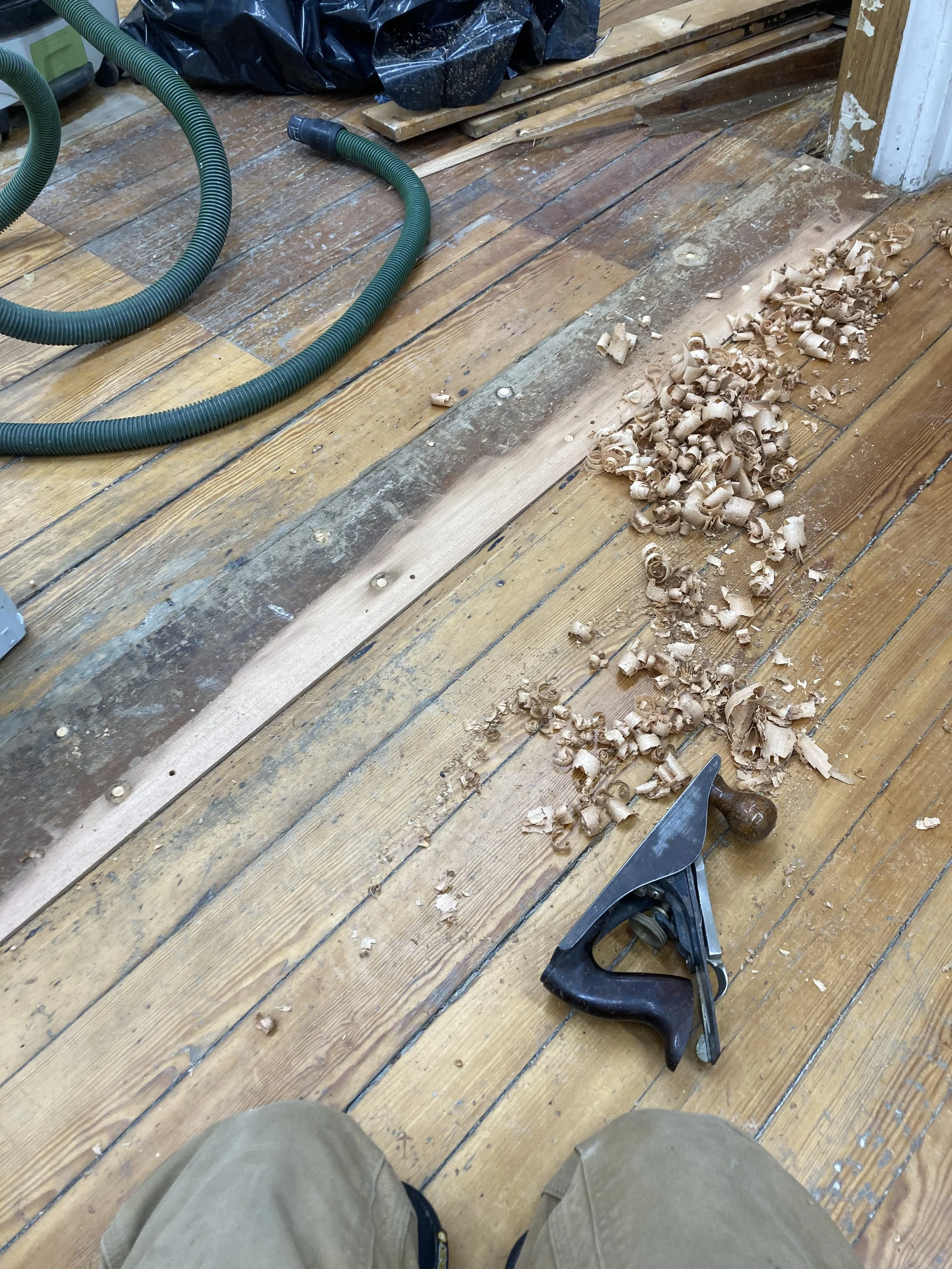 Wooden floor being sanded, shavings, hand plane, vacuum hose, and black plastic bag.