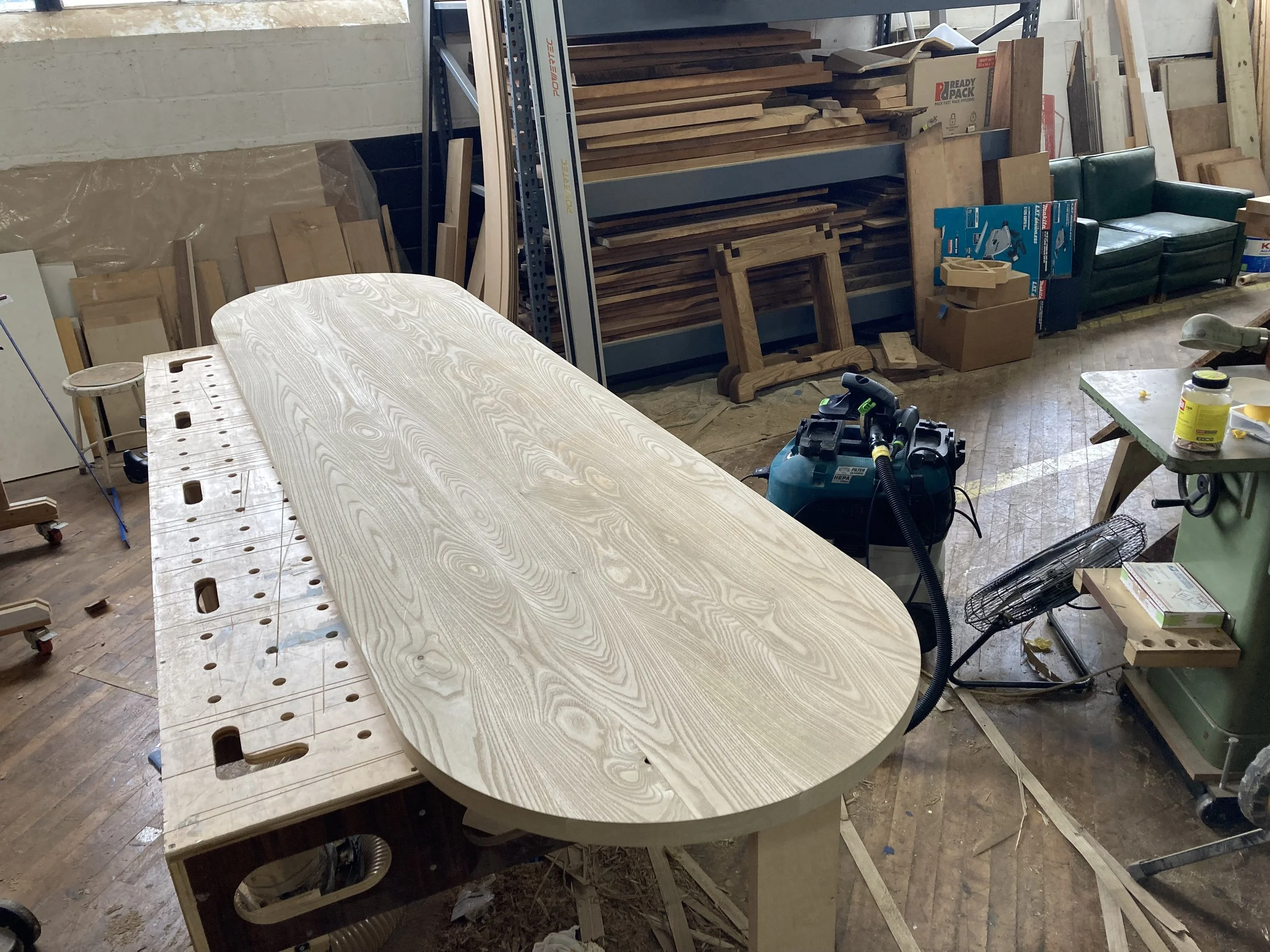Woodworking shop with an unfinished wooden table on a workbench, surrounded by tools and materials like a vacuum, fan, and various wood pieces in the background.