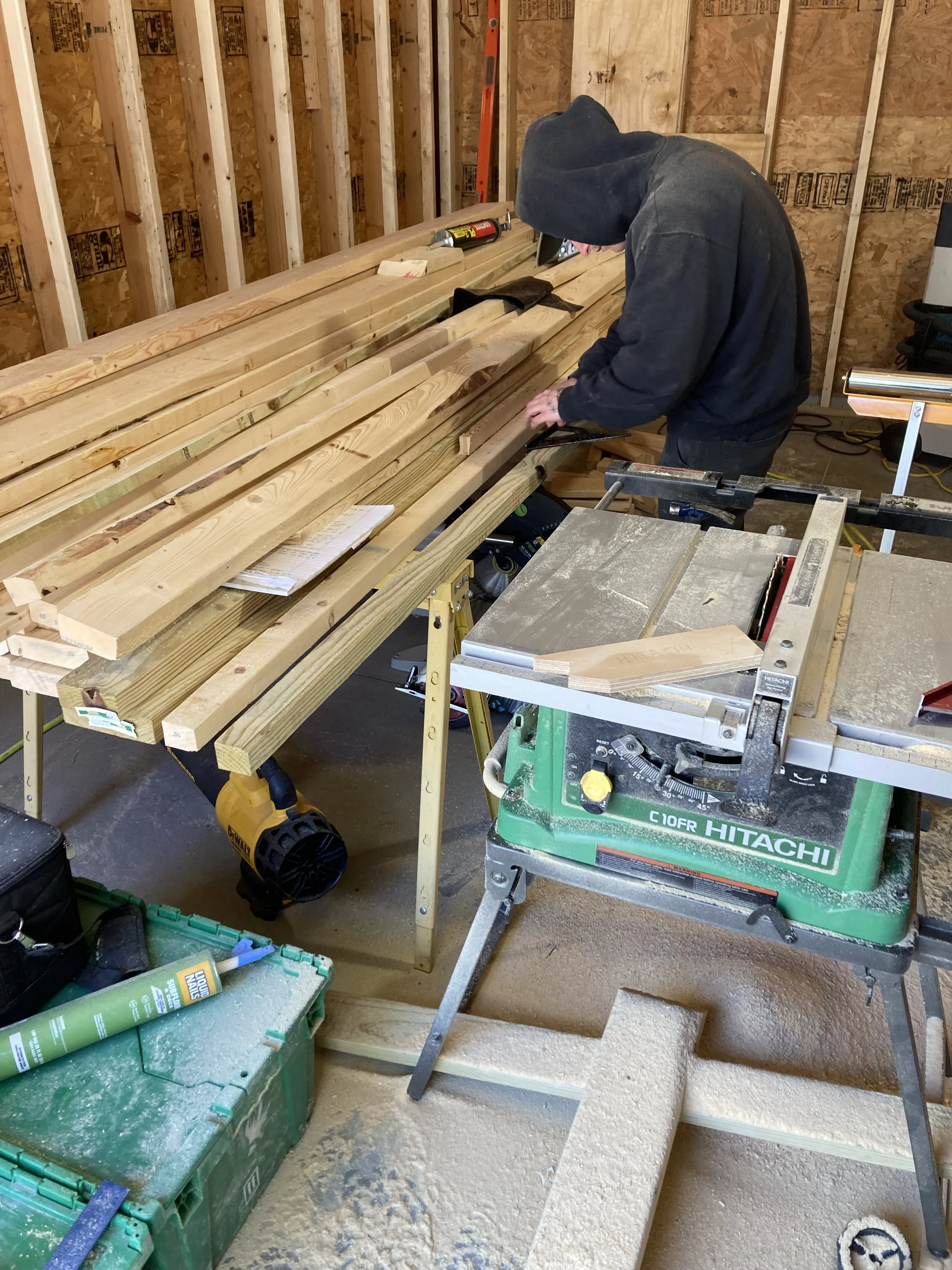 Person working in a workshop with wood planks on a table and a Hitachi table saw surrounded by sawdust and tools.