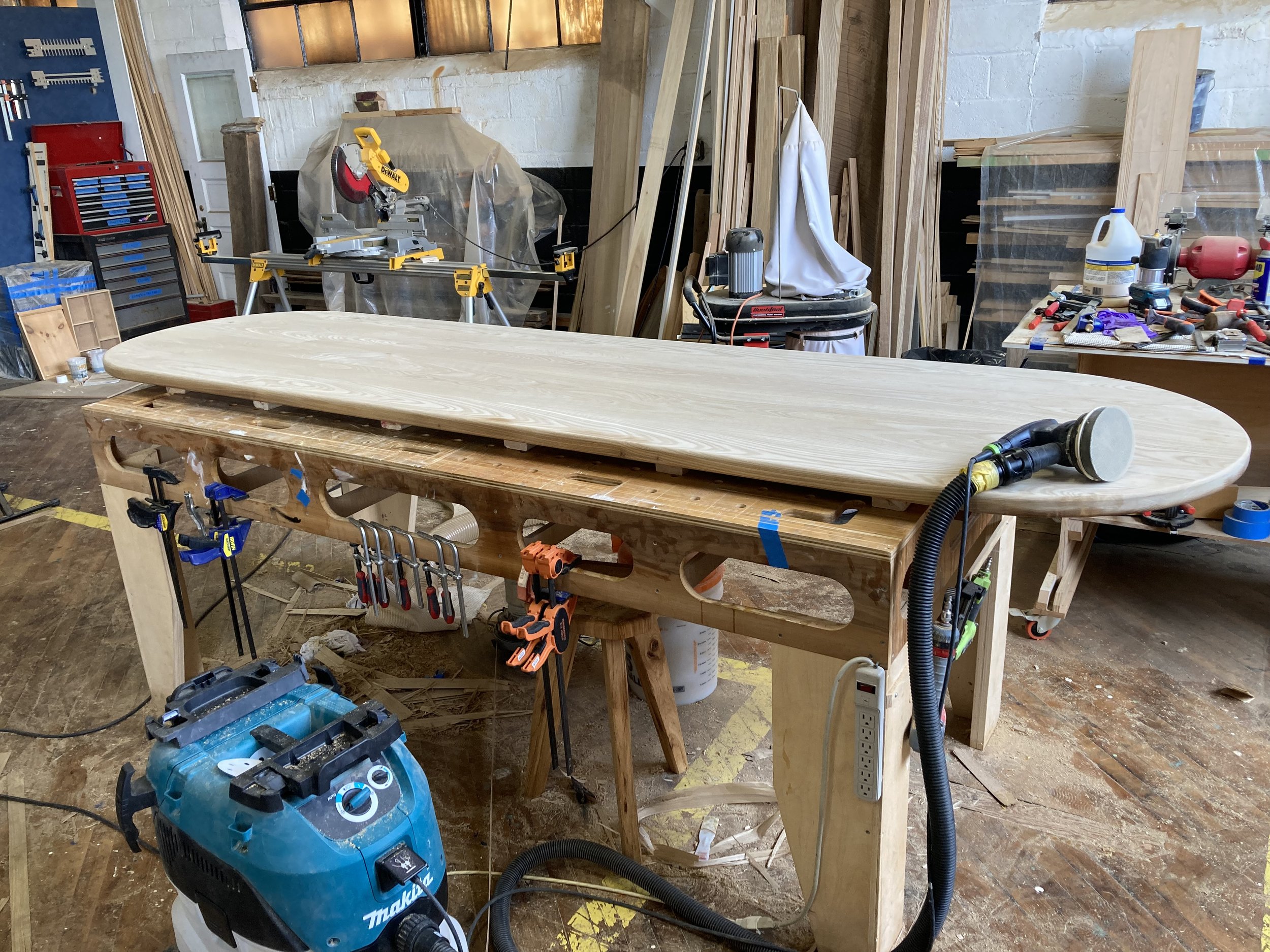 Woodworking workshop with a table partially assembled on a workbench, surrounded by tools like clamps, a miter saw, and sanding equipment.