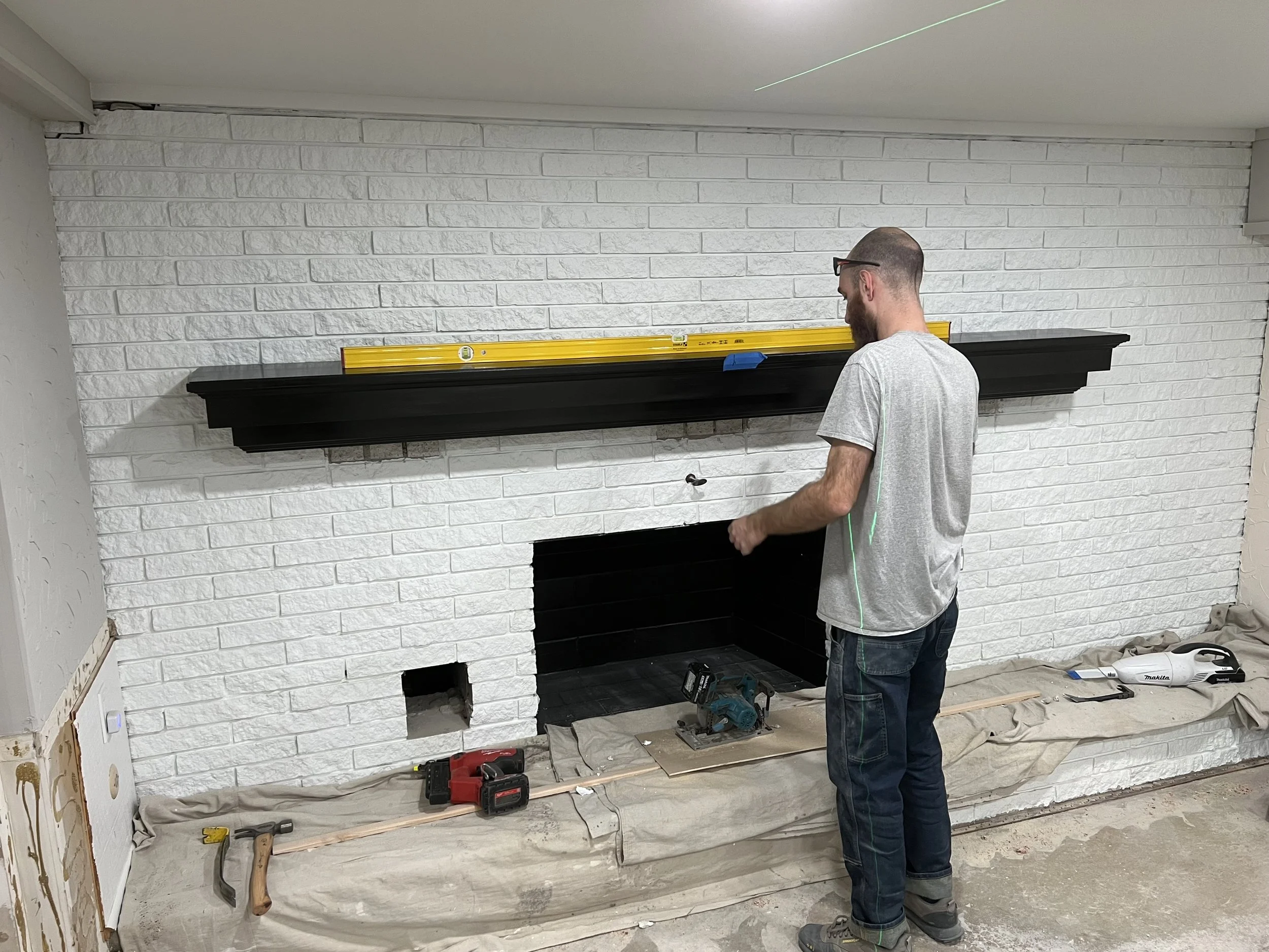 Man working on fireplace renovation with white brick wall, using a level on a dark mantel, surrounded by tools and construction materials.