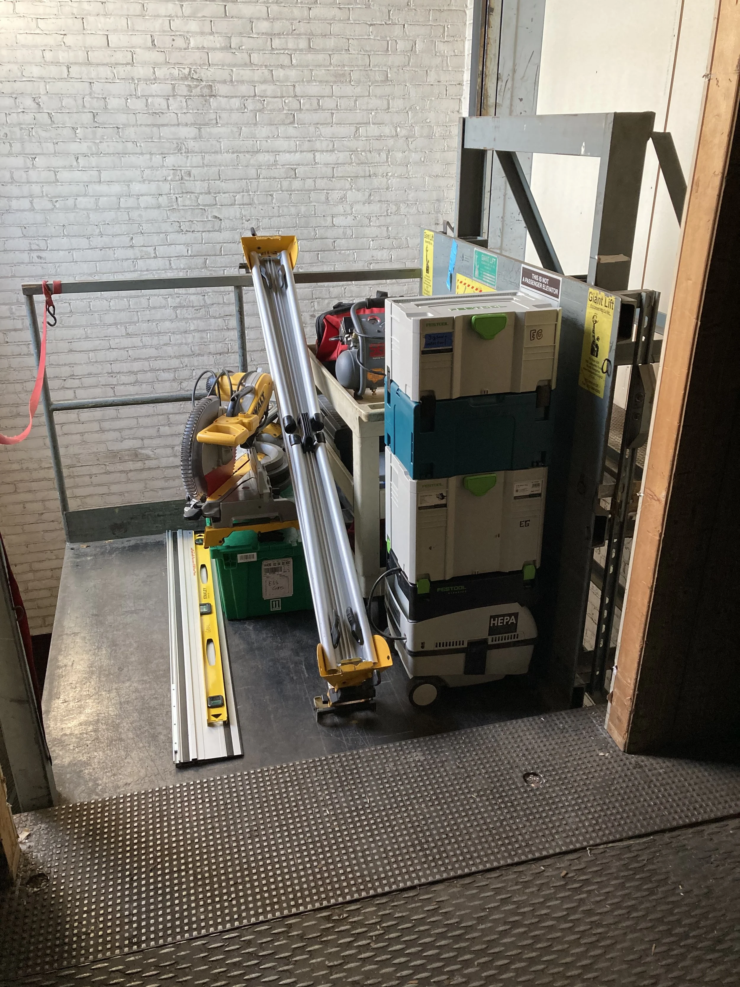 Construction tools and equipment on a lift, including a miter saw, stackable tool boxes with HEPA filter label, and a red tool bag against a white brick wall.