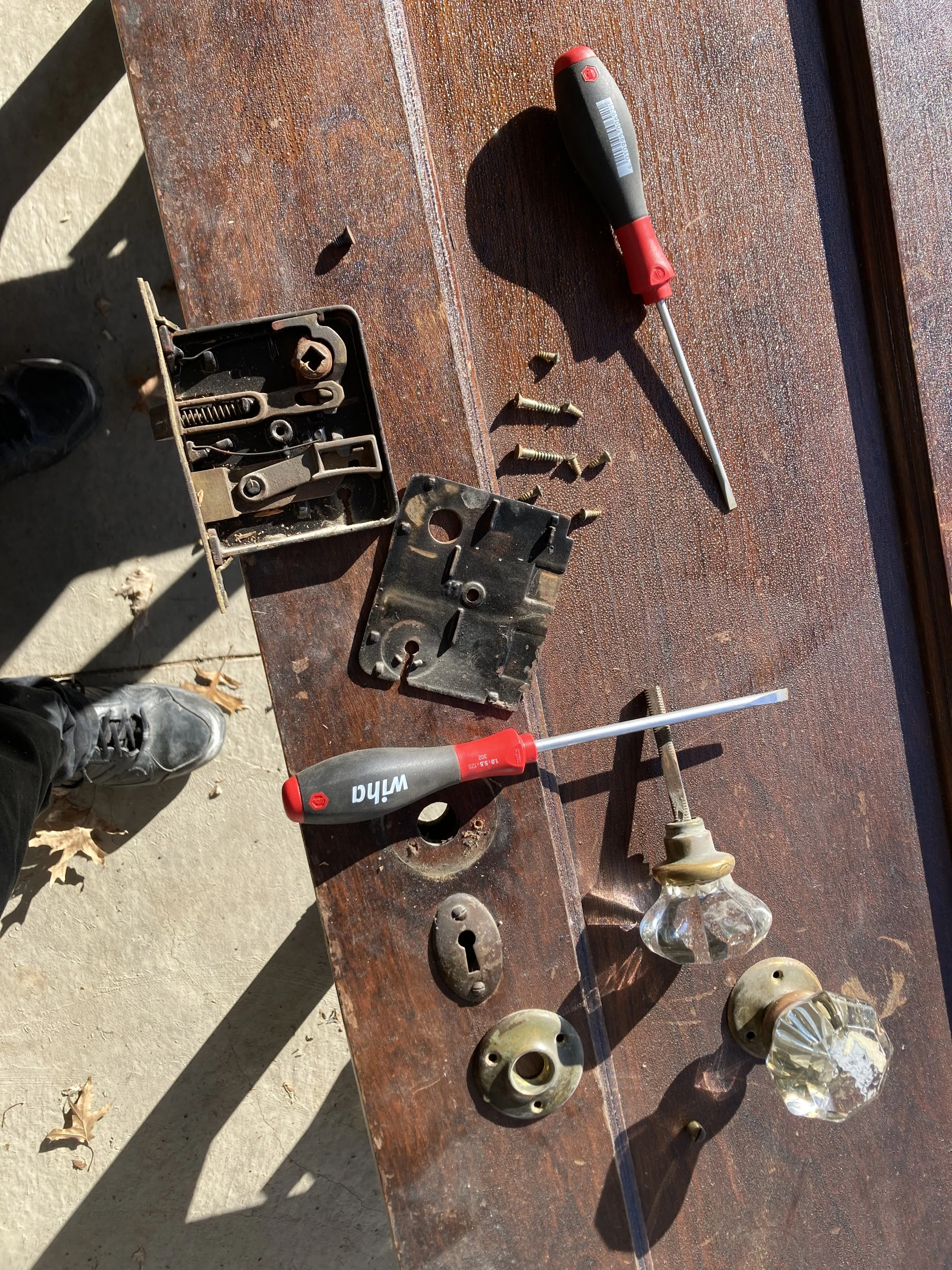 Disassembled door with crystal doorknobs, lock mechanism, screws, and two screwdrivers on a wooden surface.