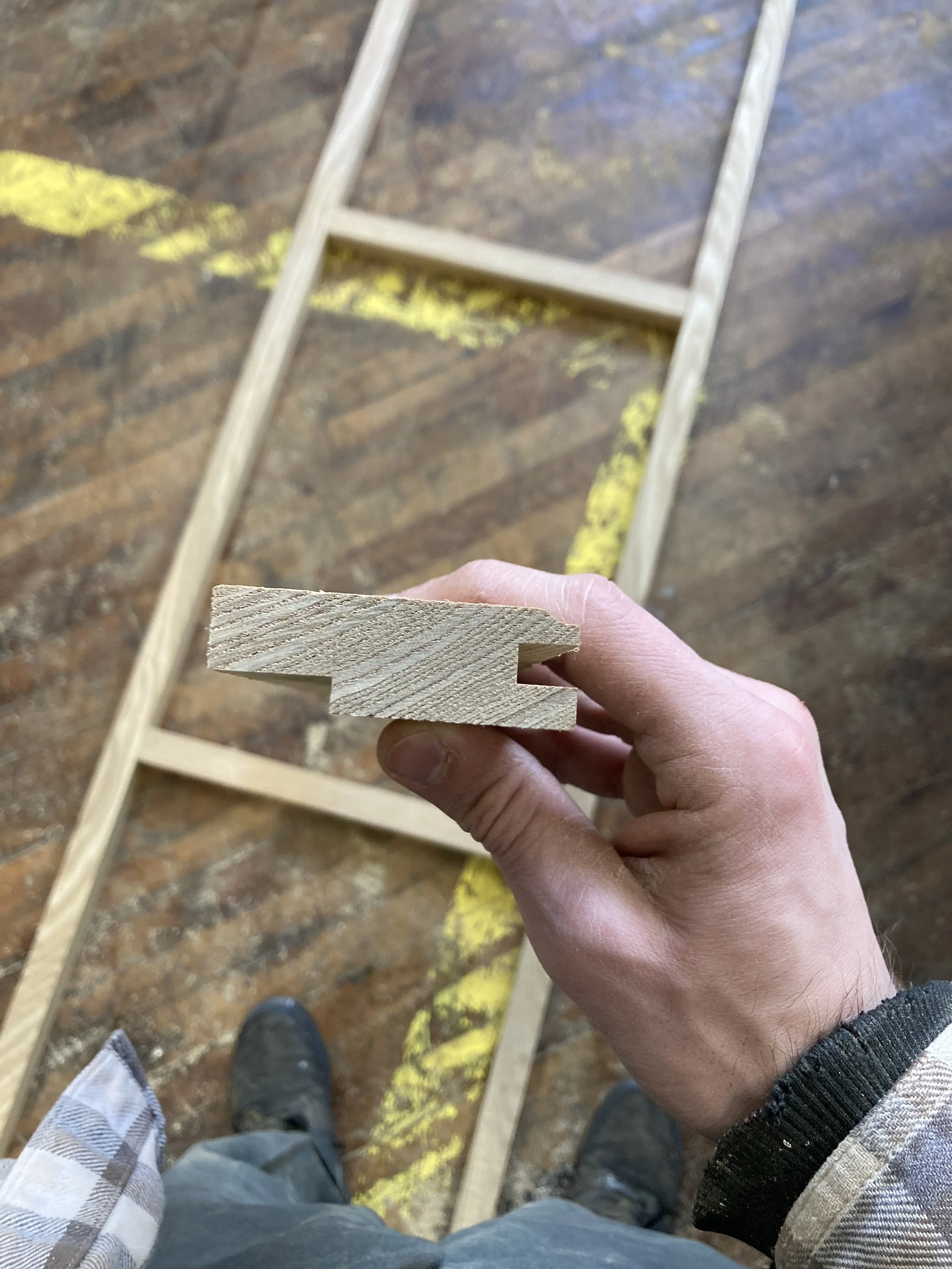 Person holding a piece of grooved wood, with a wooden frame and floor in the background.