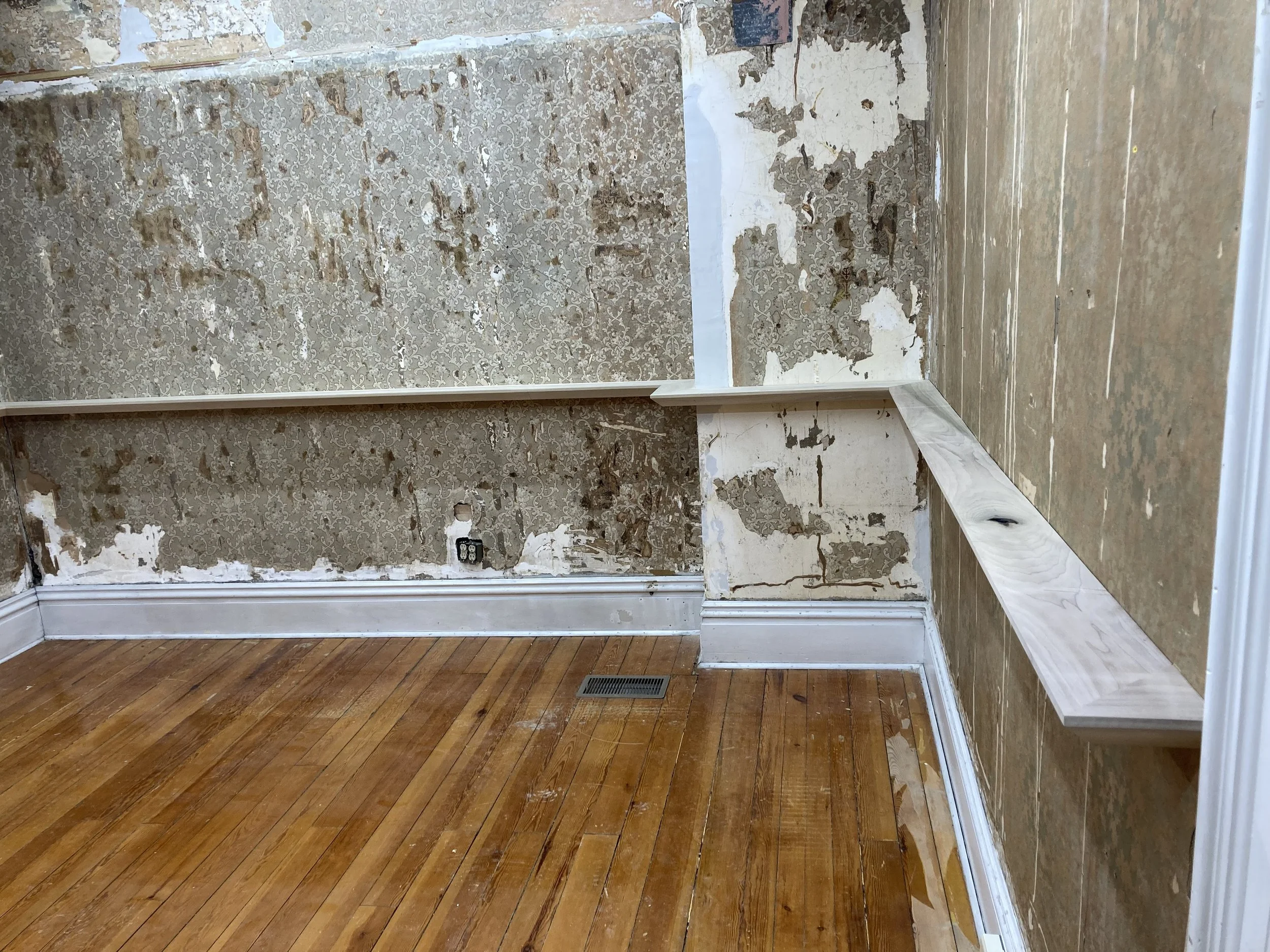 Room under renovation with peeling wallpaper and bare walls, featuring light wooden floors and newly installed narrow wooden shelves around the perimeter.