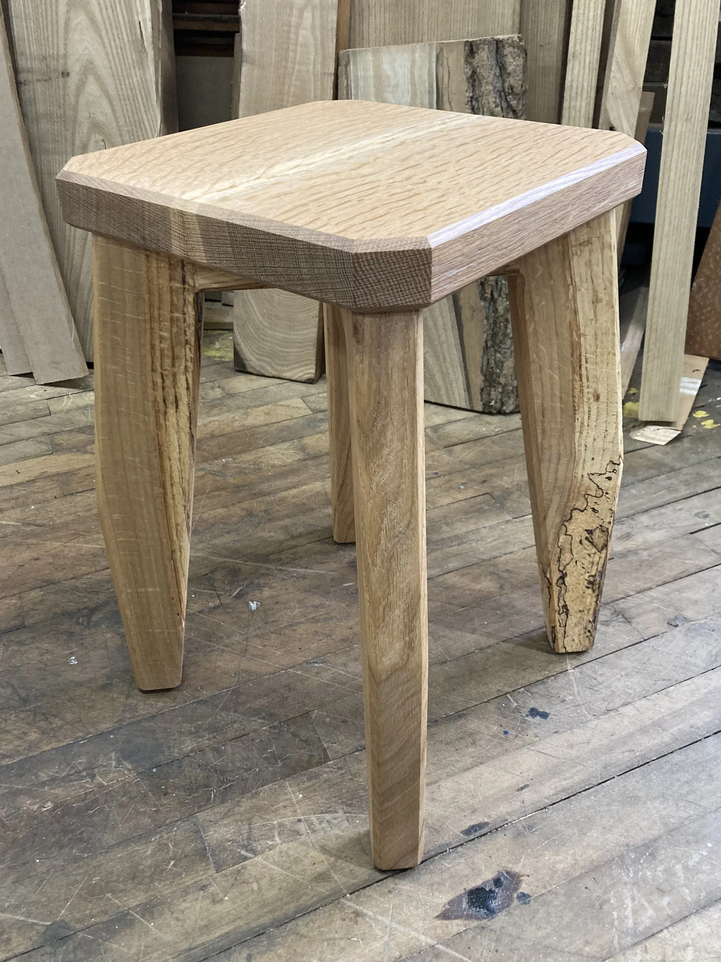 Wooden stool with four thick legs and a square seat in a workshop with wooden planks in the background.