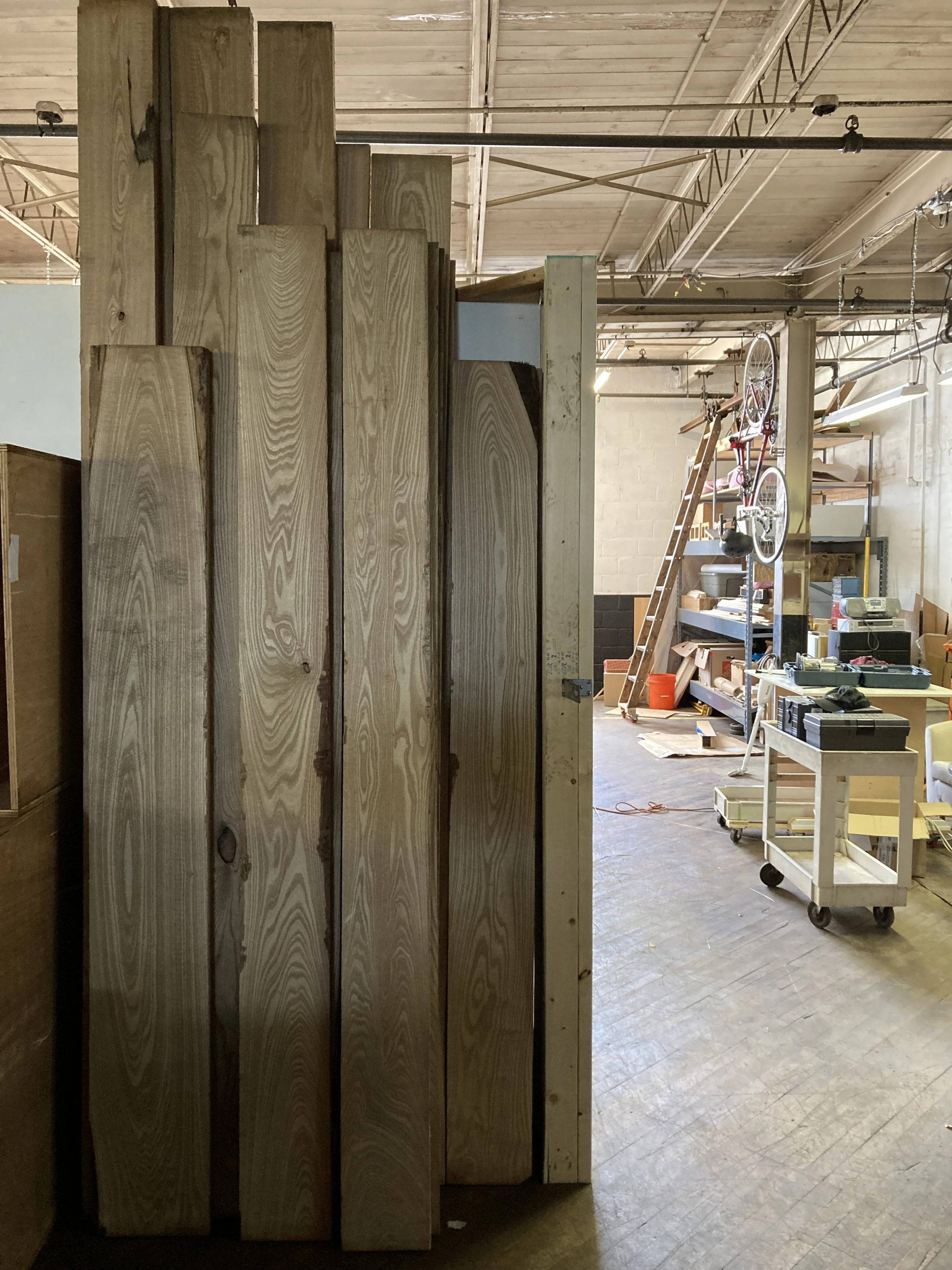 Stack of wooden planks in a workshop with tools and equipment in the background.