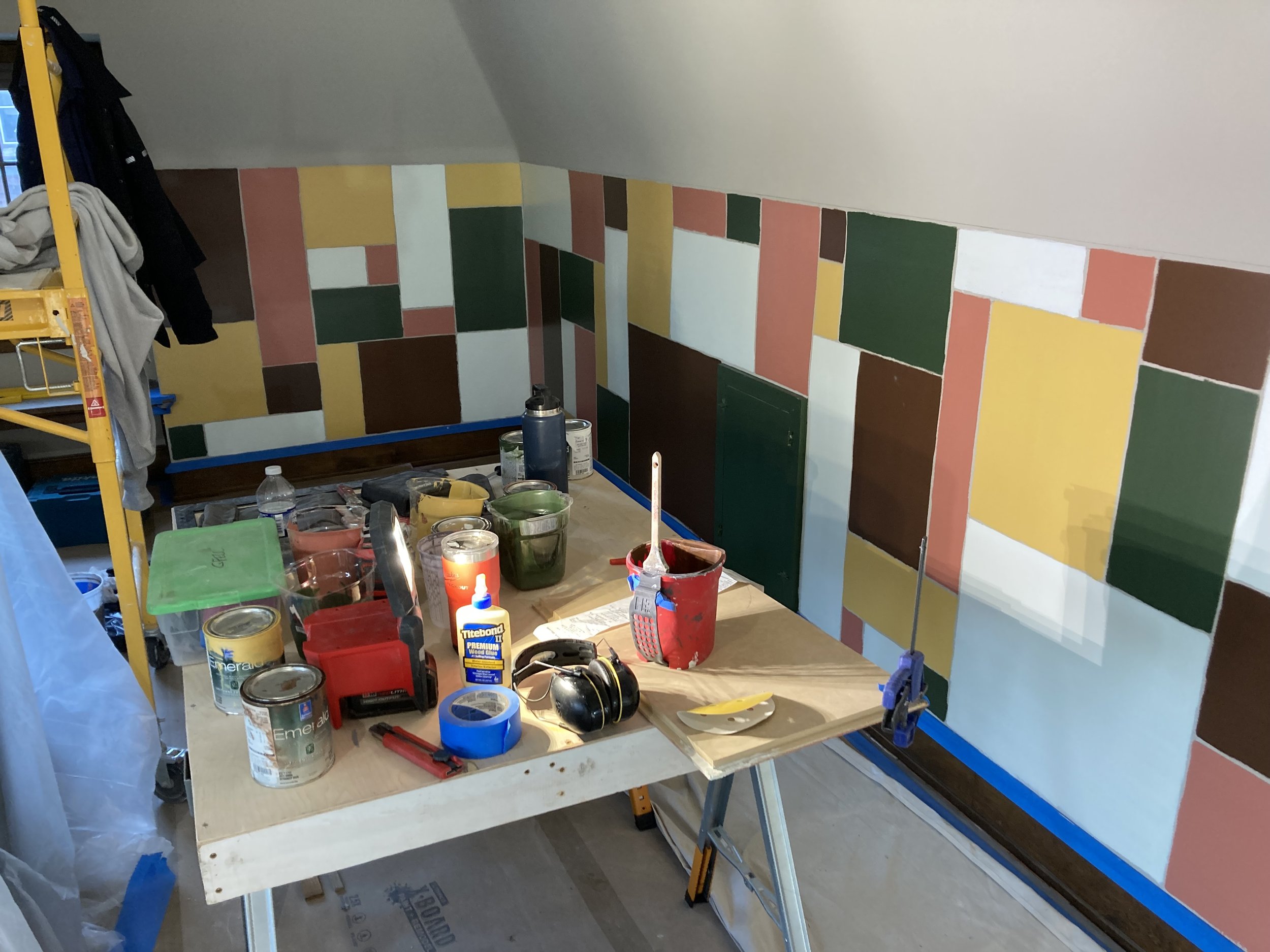 Room under renovation with a geometric multicolored wall design, tools and paint cans on a wooden table, and a scaffold with clothes hanging nearby.