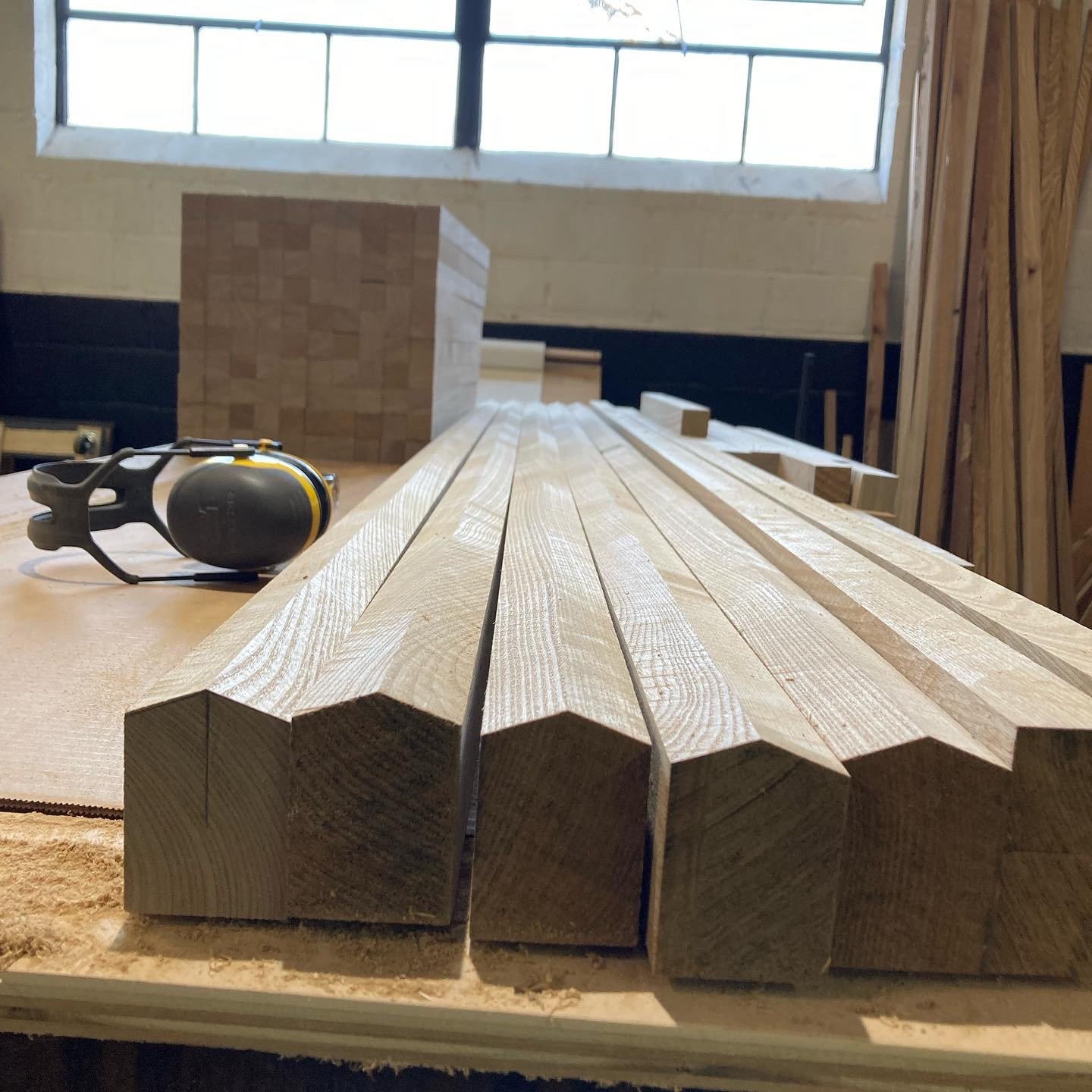 Wooden beams on a workbench next to safety earmuffs, with a block structure in the background and window light.