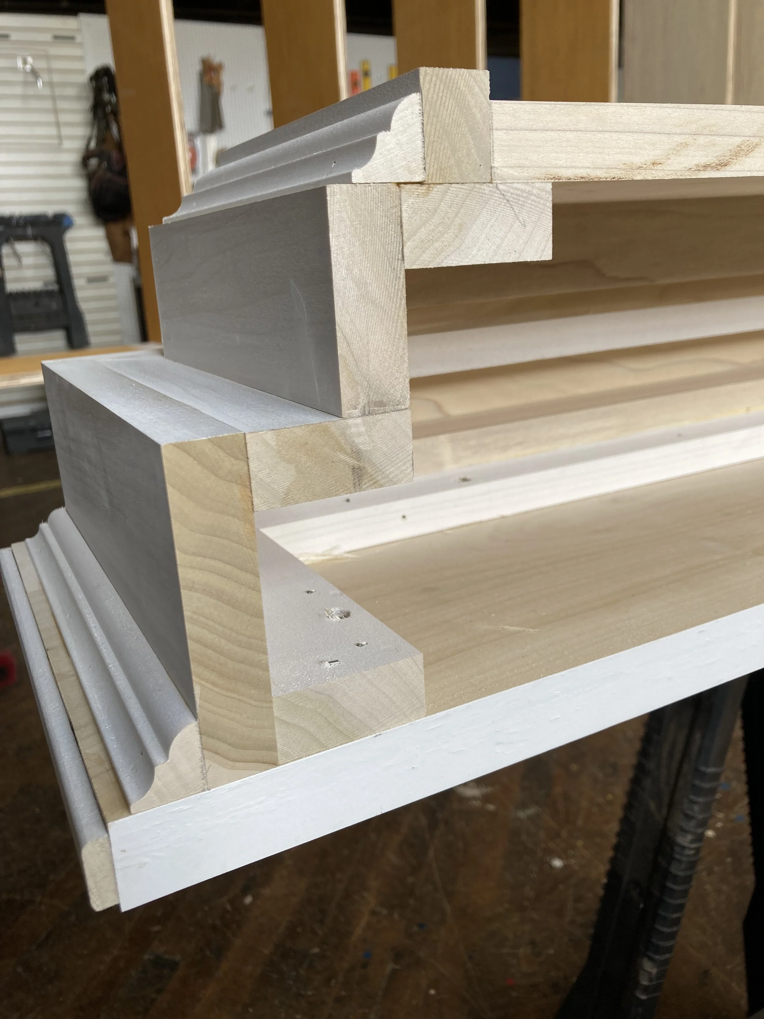 Close-up of wooden trim pieces in a workshop, arranged to form a corner frame, with visible layered joints and molding details.