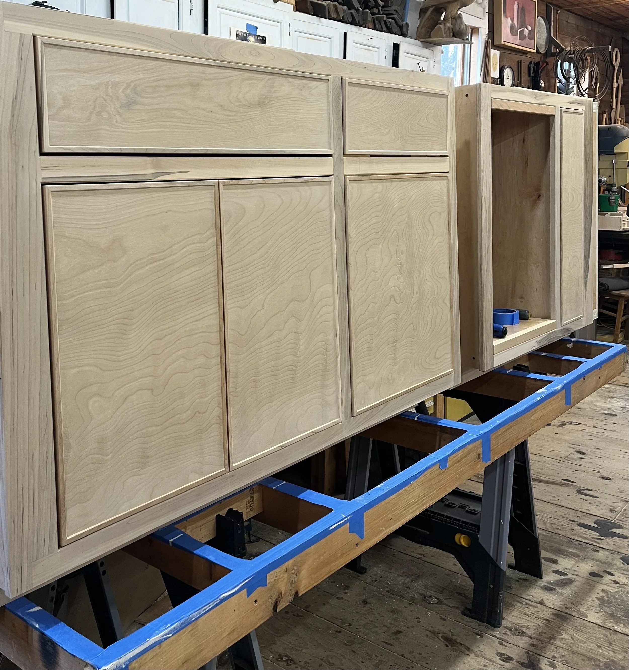 Wooden cabinetry in a workshop under construction, with unfinished doors and drawers, placed on a workbench with blue tape markings.