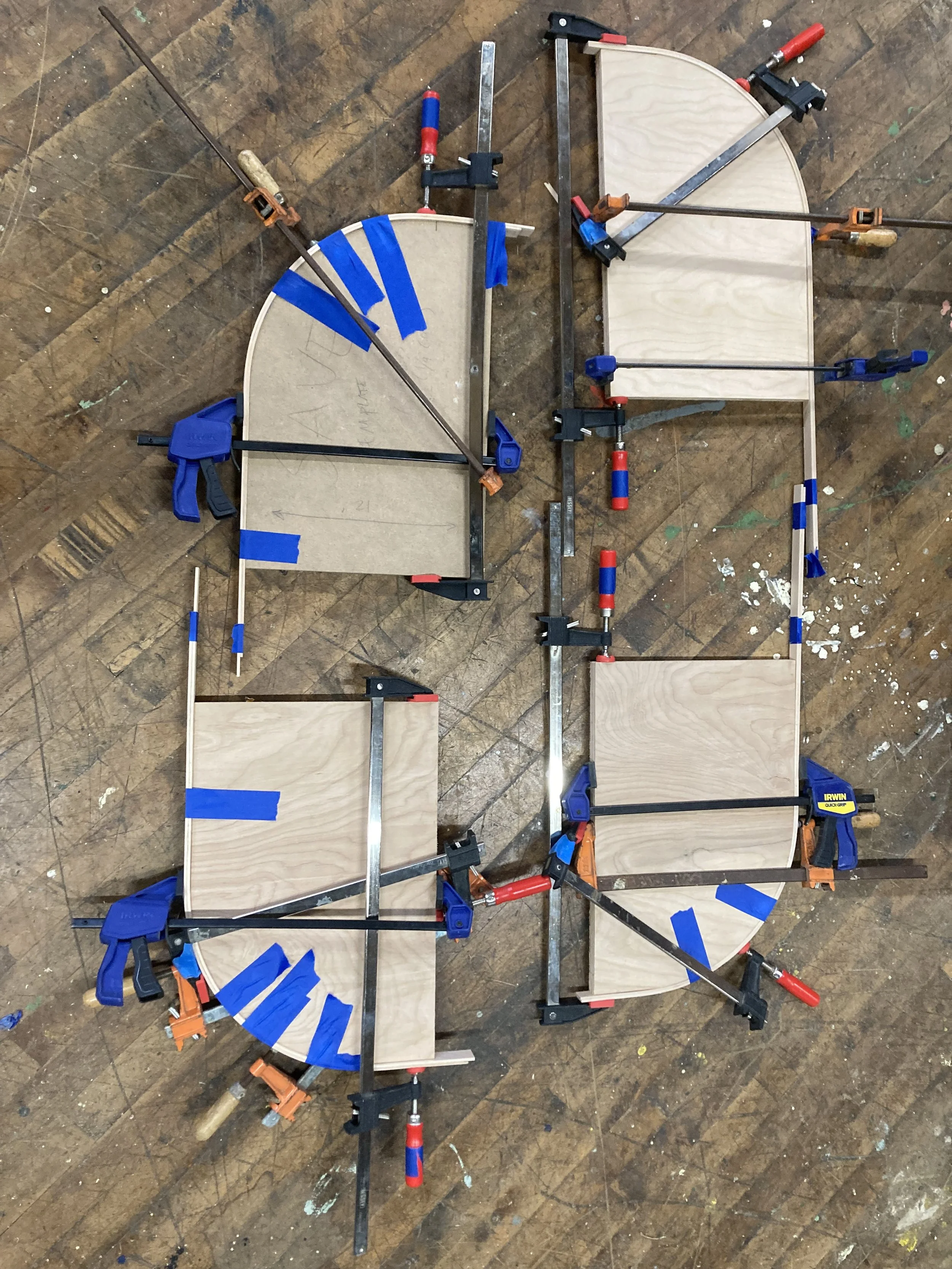 Woodwork pieces clamped together with various clamps, some blue painter's tape on the pieces, on a workshop floor.