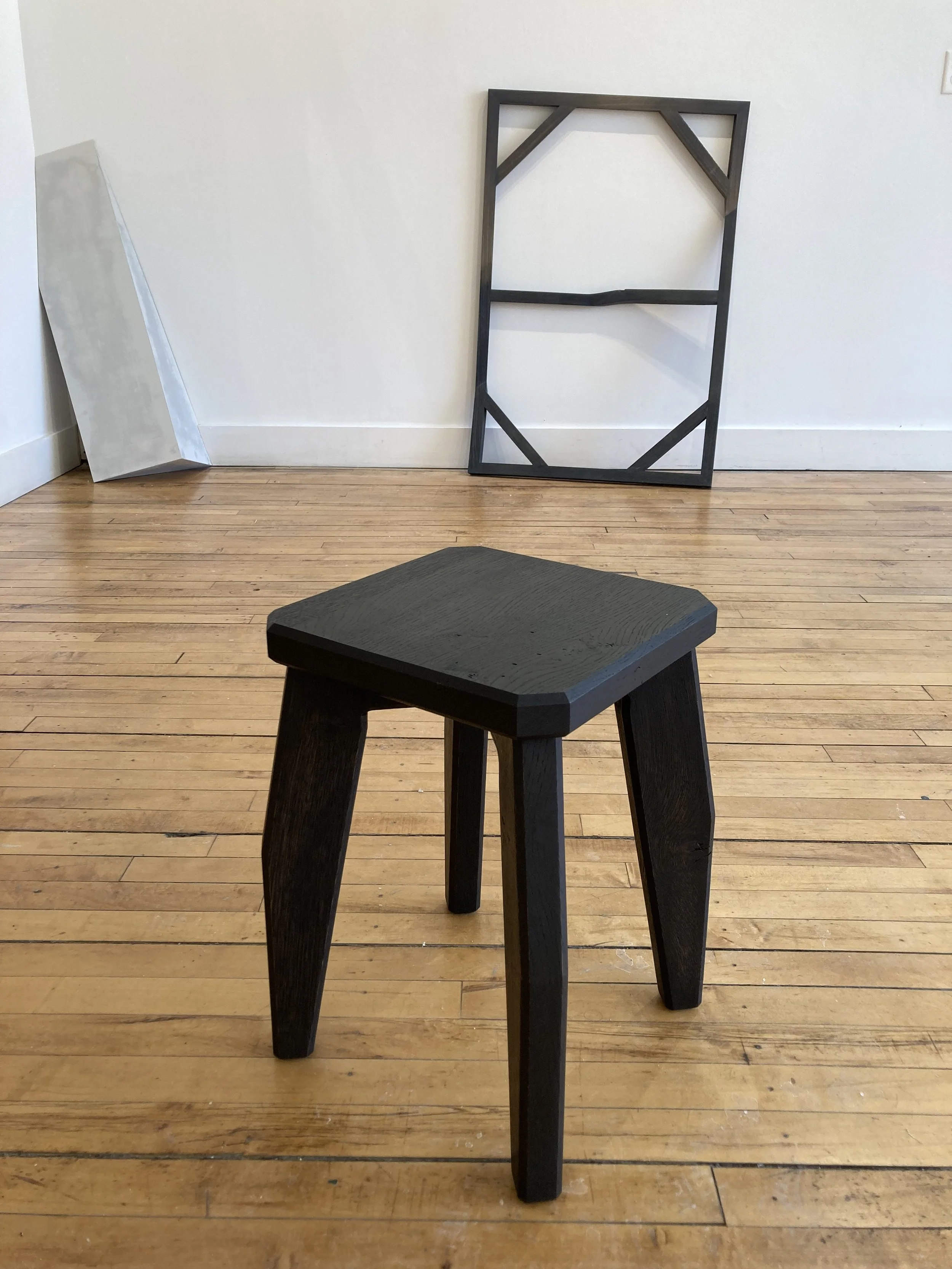 A small wooden stool on a hardwood floor with a leaning empty frame and a canvas against a white wall.