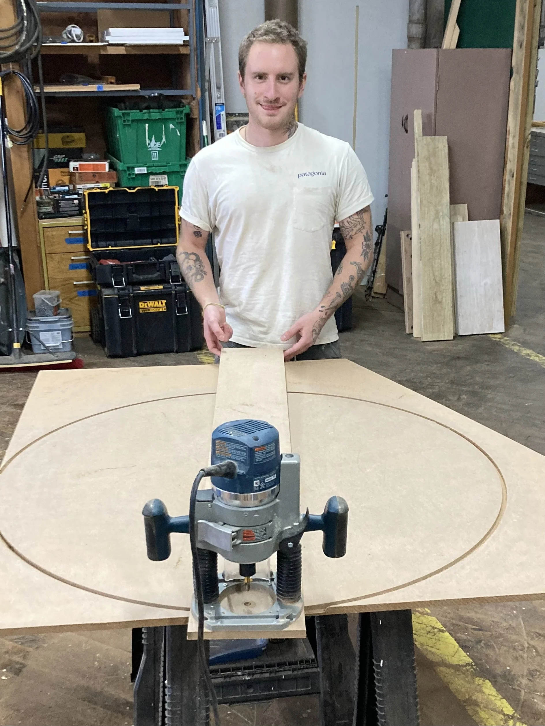 Person in a workshop using a router to cut a circle in a wooden board, surrounded by tools and materials.