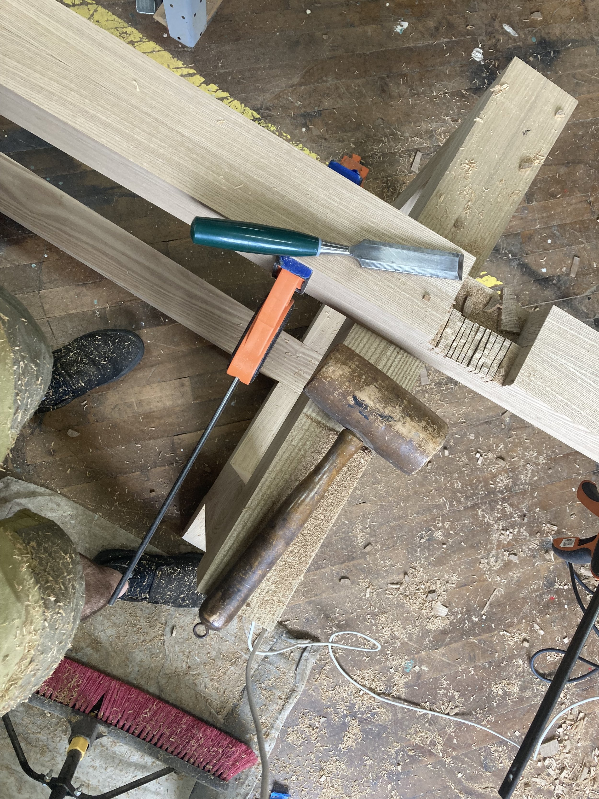 Woodworking setup with wooden beams, a chisel, a mallet, a clamp, a saw, and sawdust on the floor.