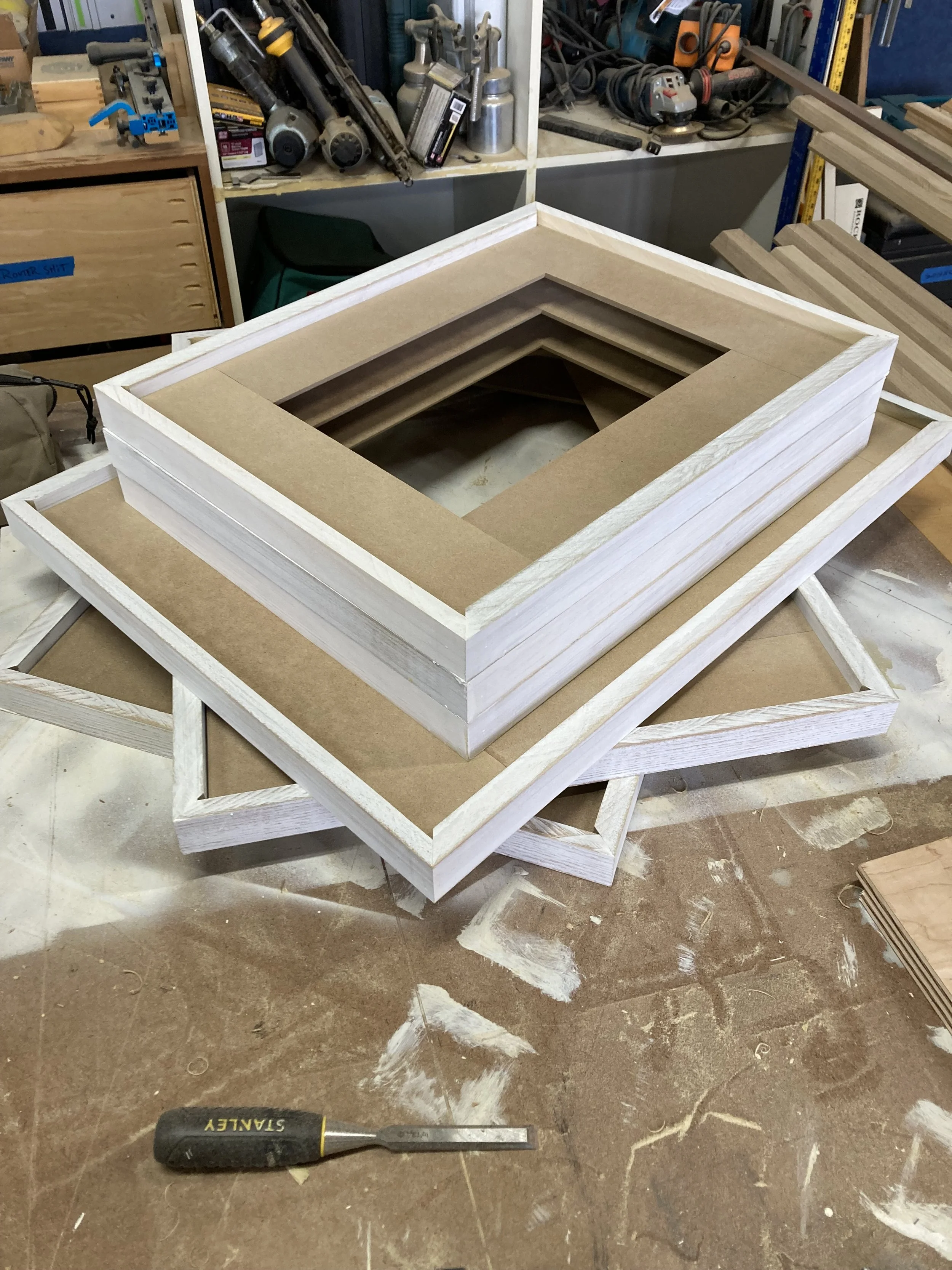 A stack of unfinished wooden picture frames on a workbench in a woodworking workshop with tools and equipment in the background.