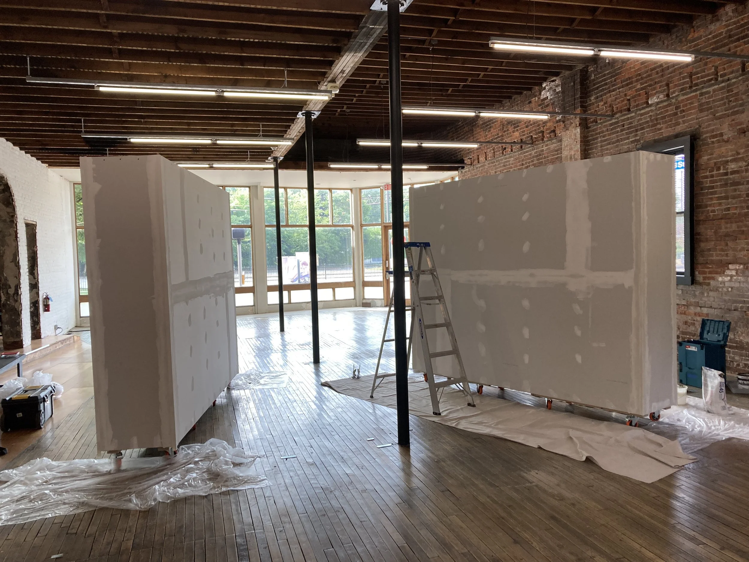 Interior of a room with two large drywall partitions, a ladder, exposed brick walls, wooden floors, and large windows. Construction materials are scattered around.