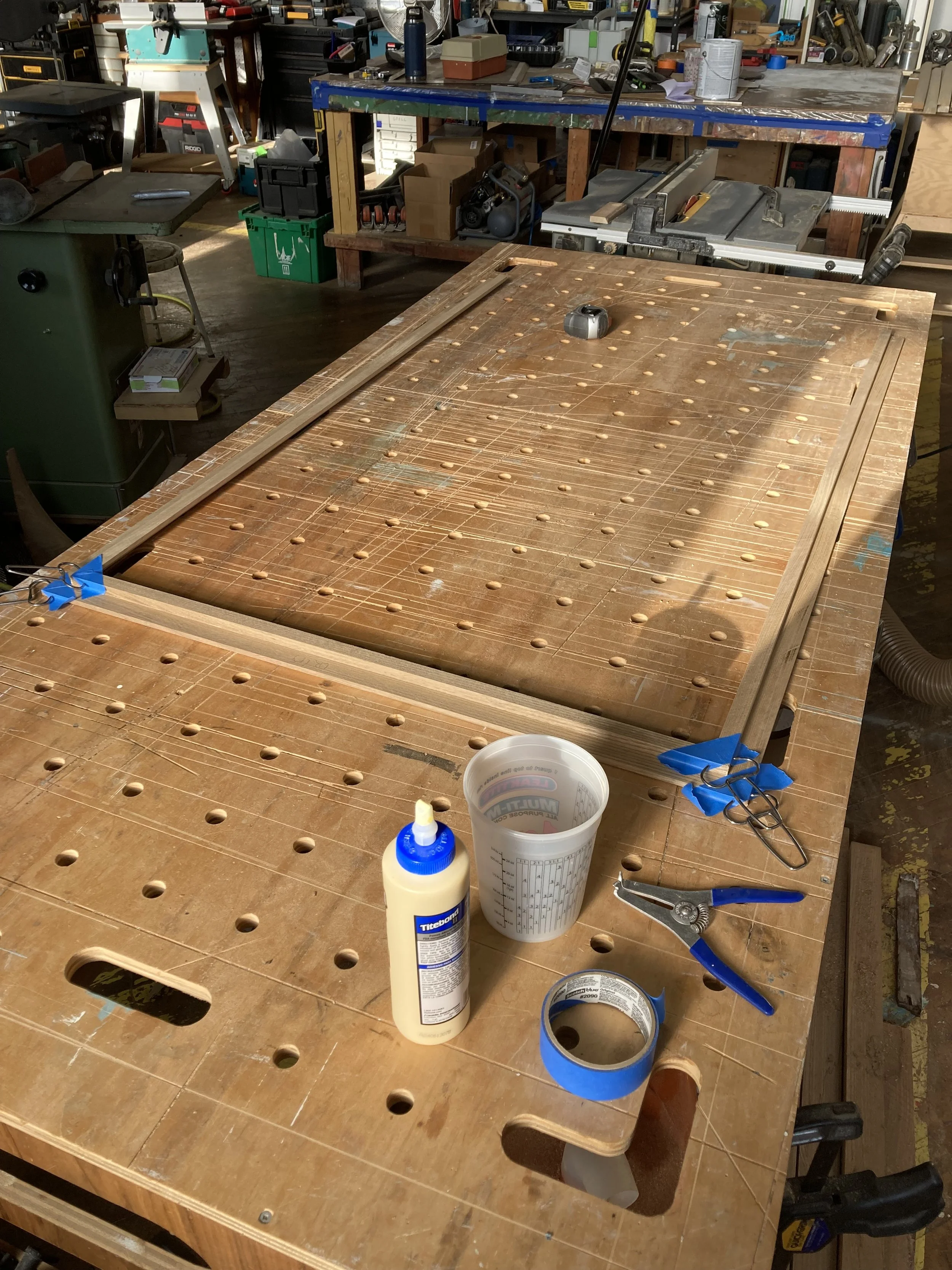 Woodworking table with glue, measuring cup, tape, and tools in a workshop setting.