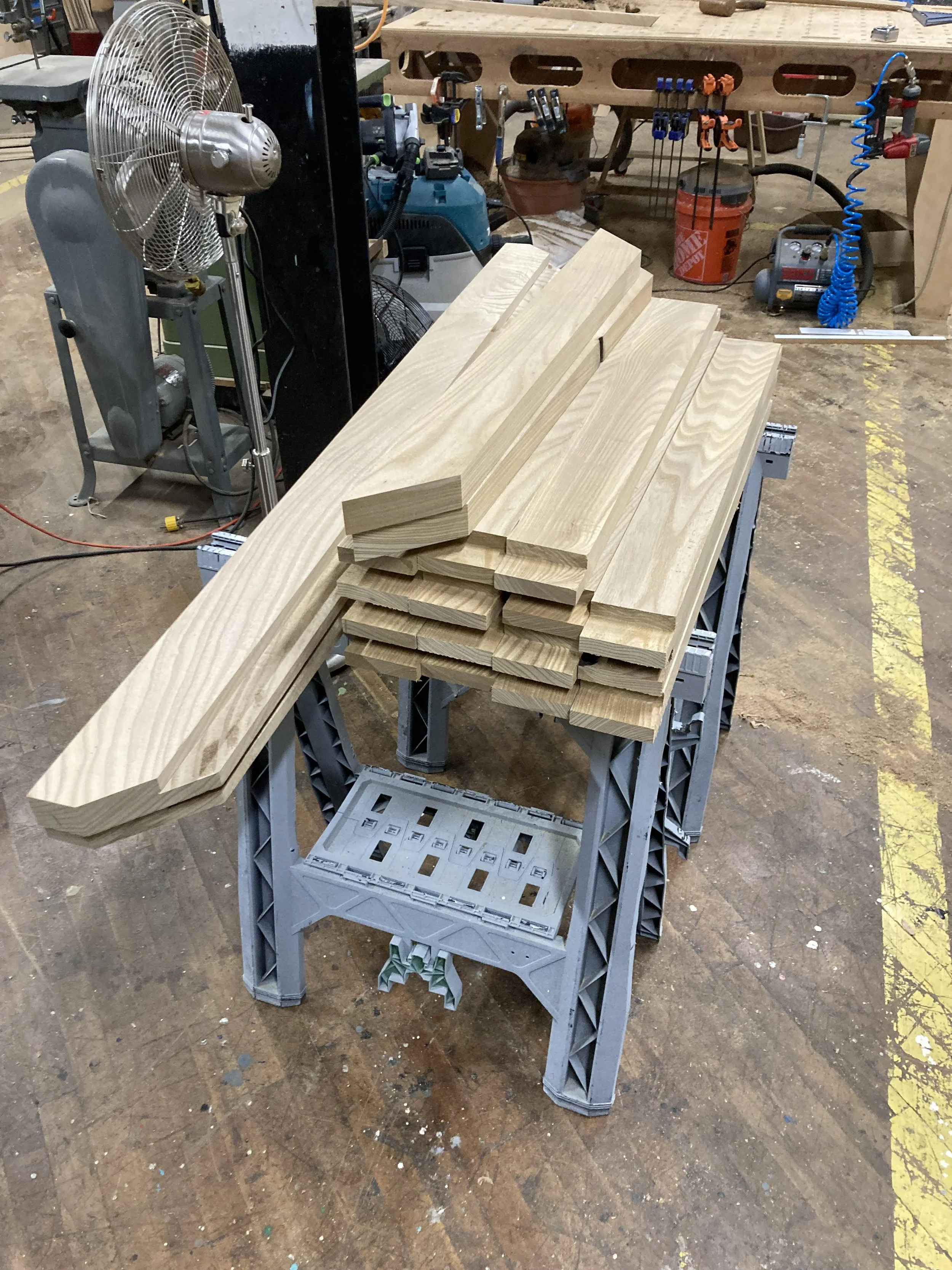 Wooden planks stacked on a sawhorse in a workshop with tools and equipment in the background.