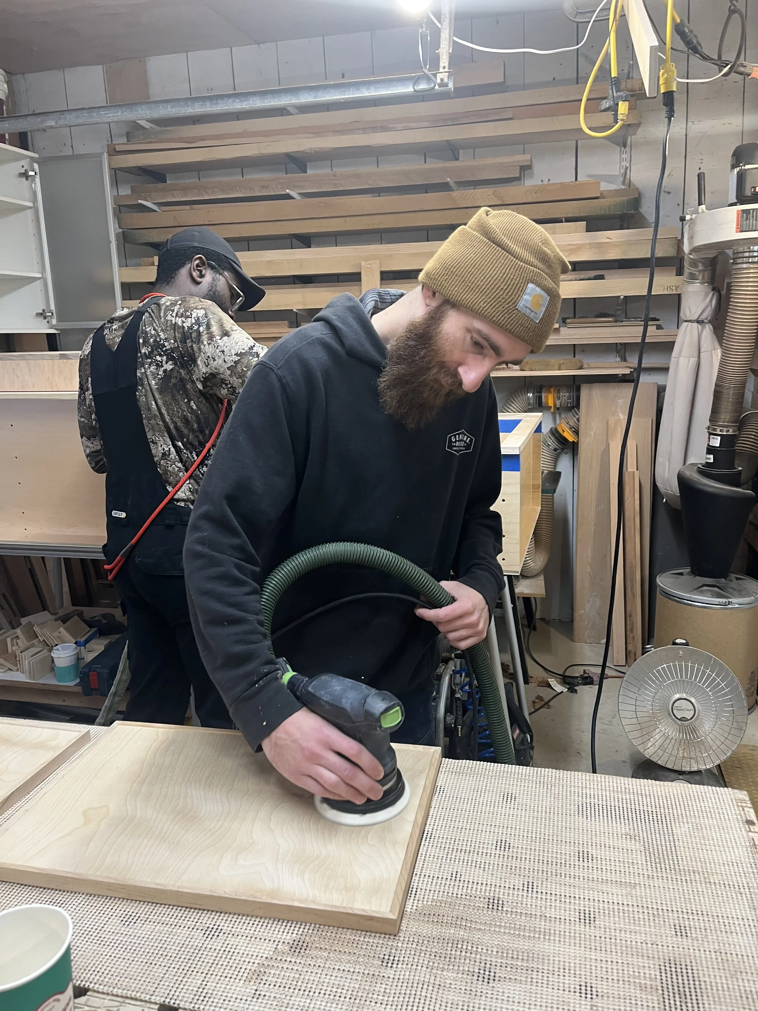Two people working in a woodworking shop, one using a sander on a wood panel.