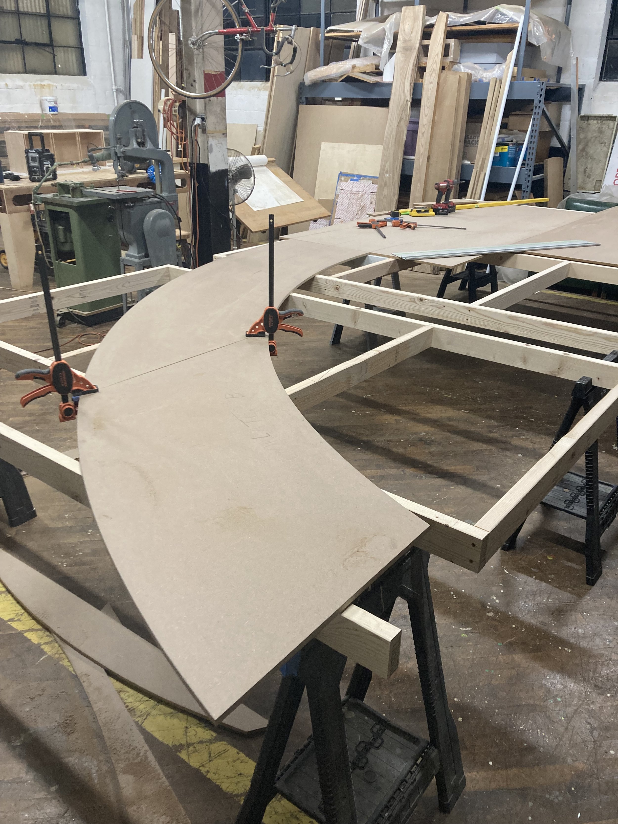 Woodworking workshop with a curved wooden structure on a frame, clamped and surrounded by tools and machinery.