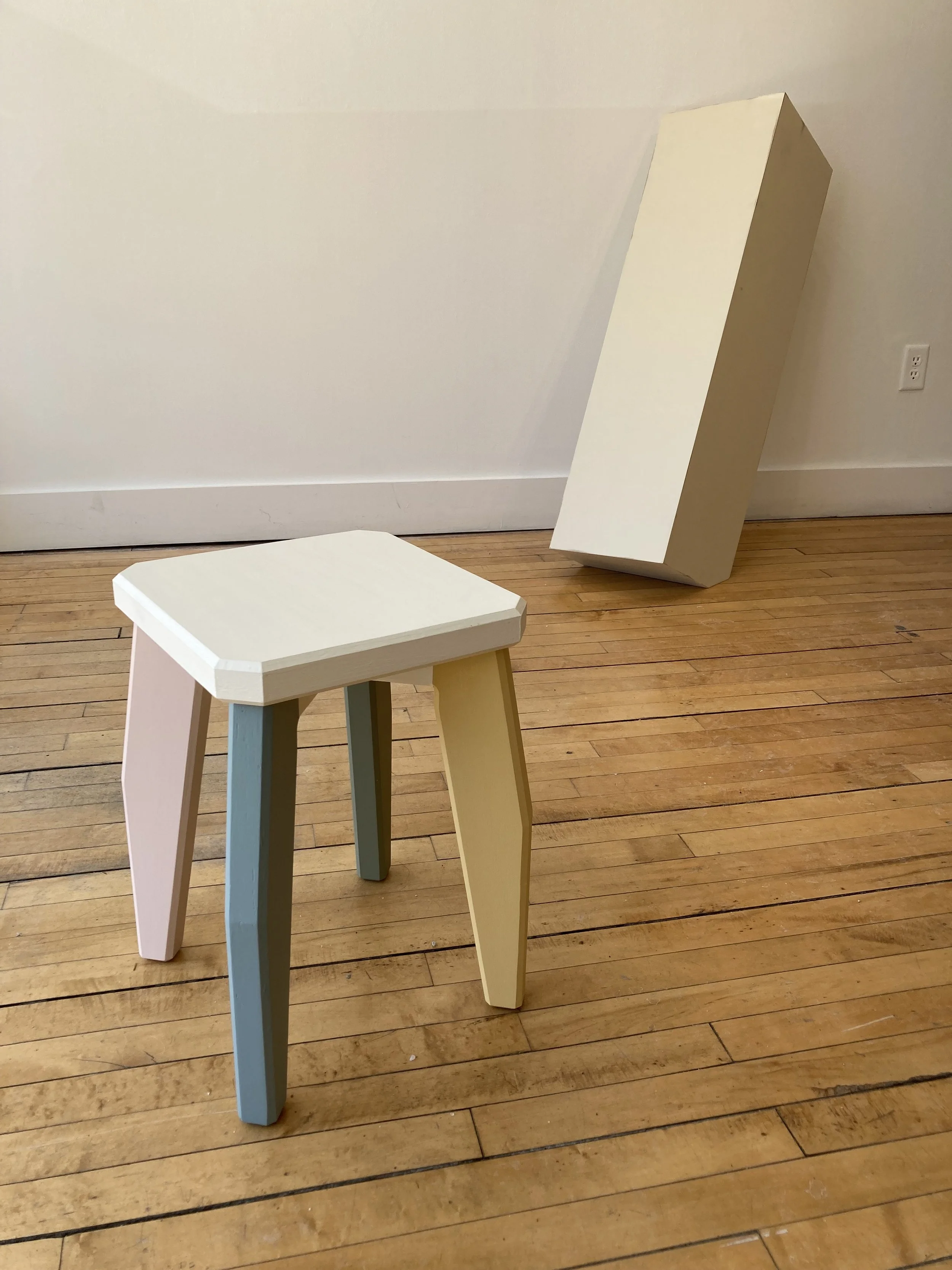 A small stool with pastel-colored legs on a wooden floor, next to a tall rectangular object leaning against a wall.