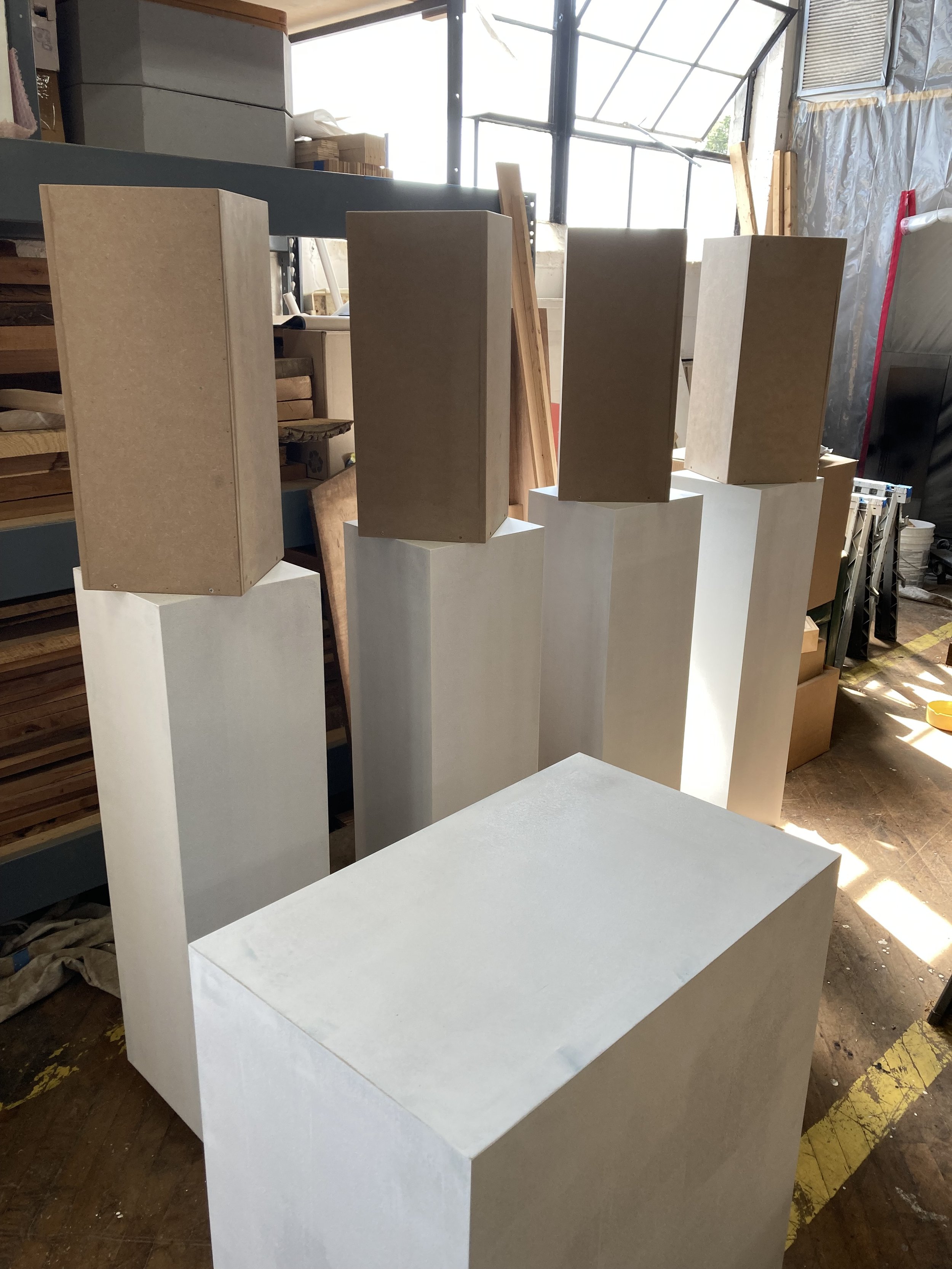 Several rectangular white and beige pedestals in a workshop setting, with various materials and tools in the background.