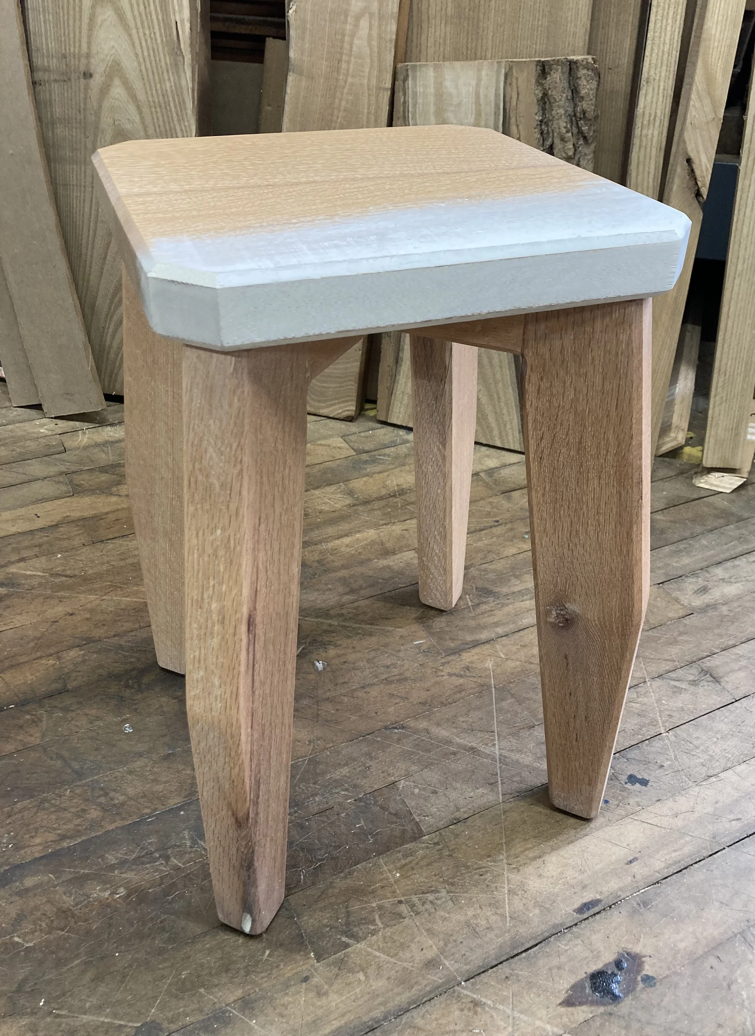 Wooden stool with unfinished seat and legs on a wooden floor in a workshop.