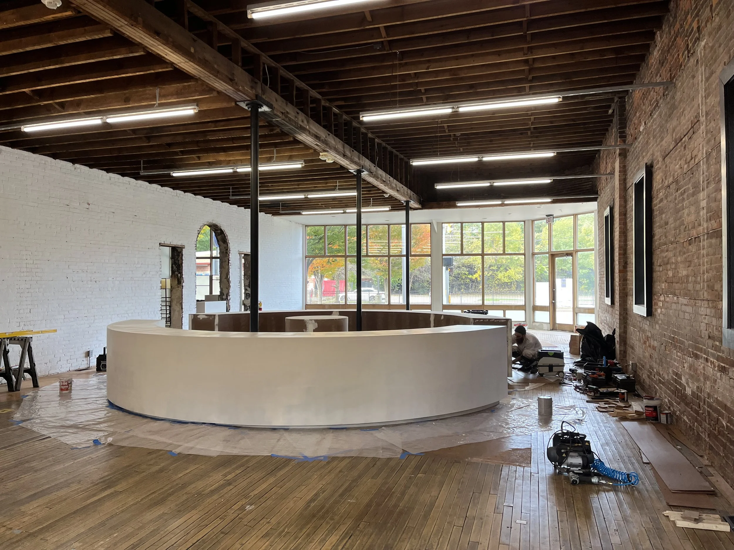 Interior of a partially renovated room with a circular white counter, wooden floors, exposed brick walls, exposed beams, and large windows.