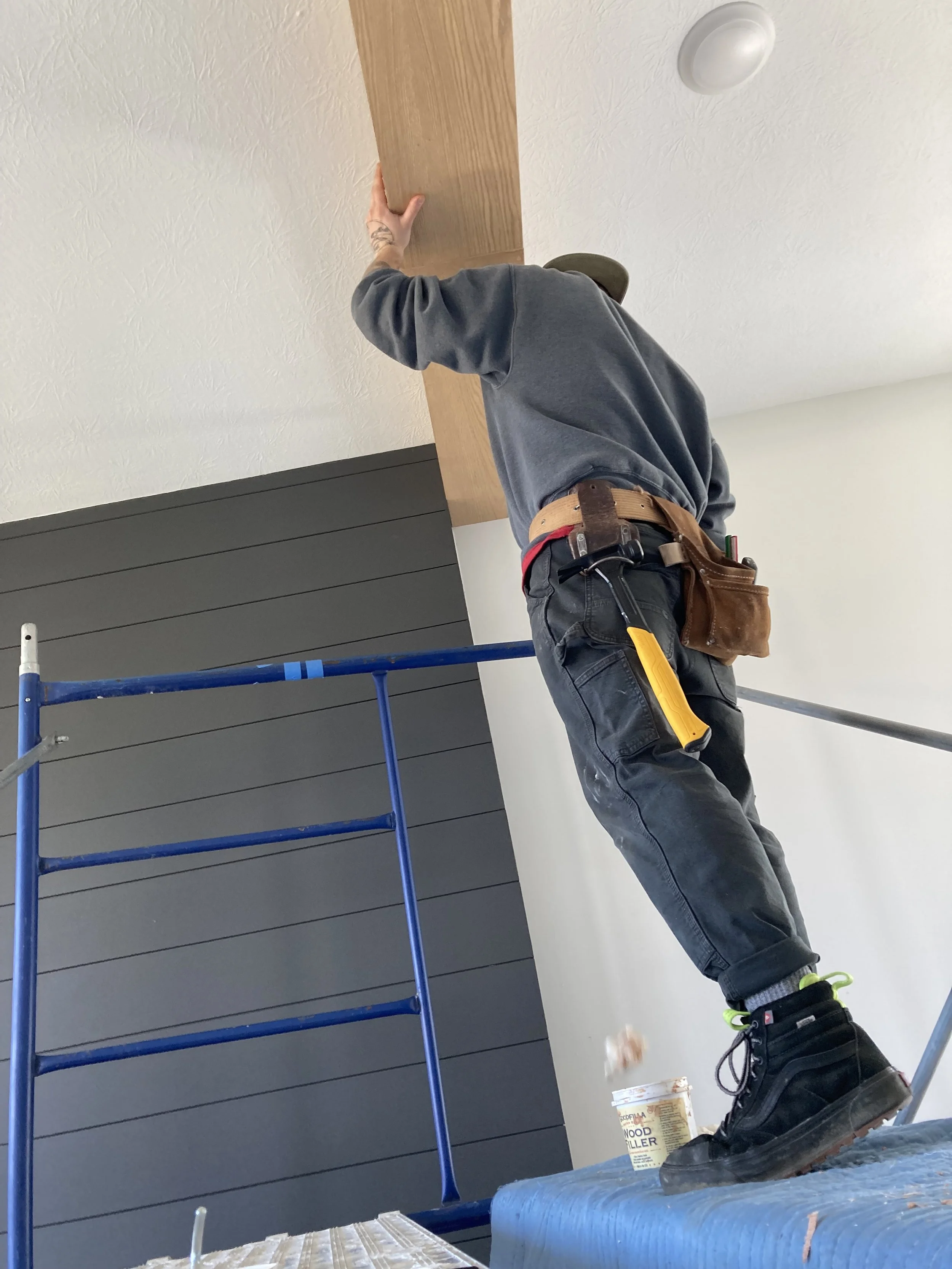 Person on scaffolding installing or adjusting a wooden beam on the ceiling, wearing a tool belt and safety shoes.