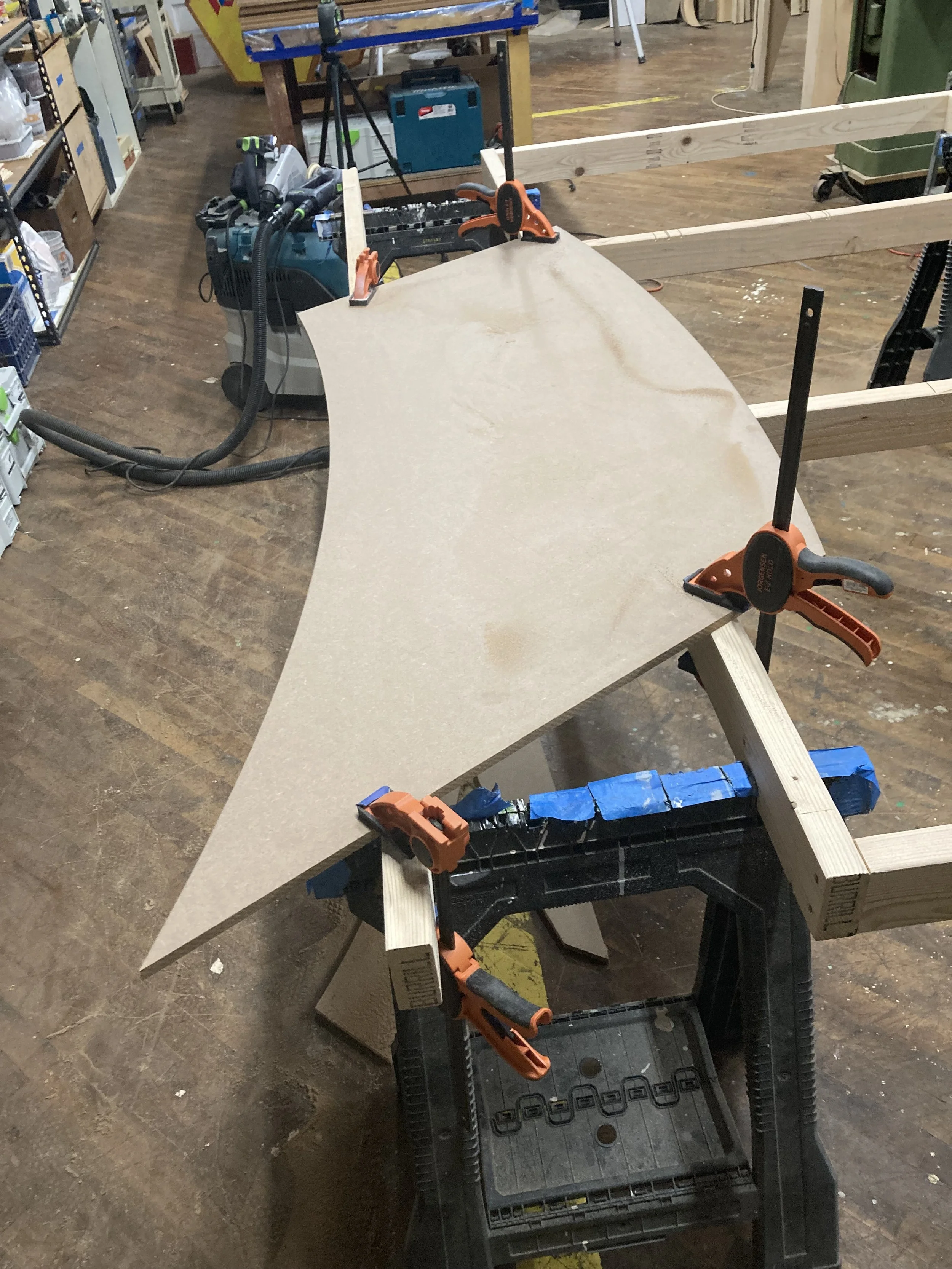 Woodworking setup with a curved wooden panel secured by clamps on a sawhorse in a workshop.